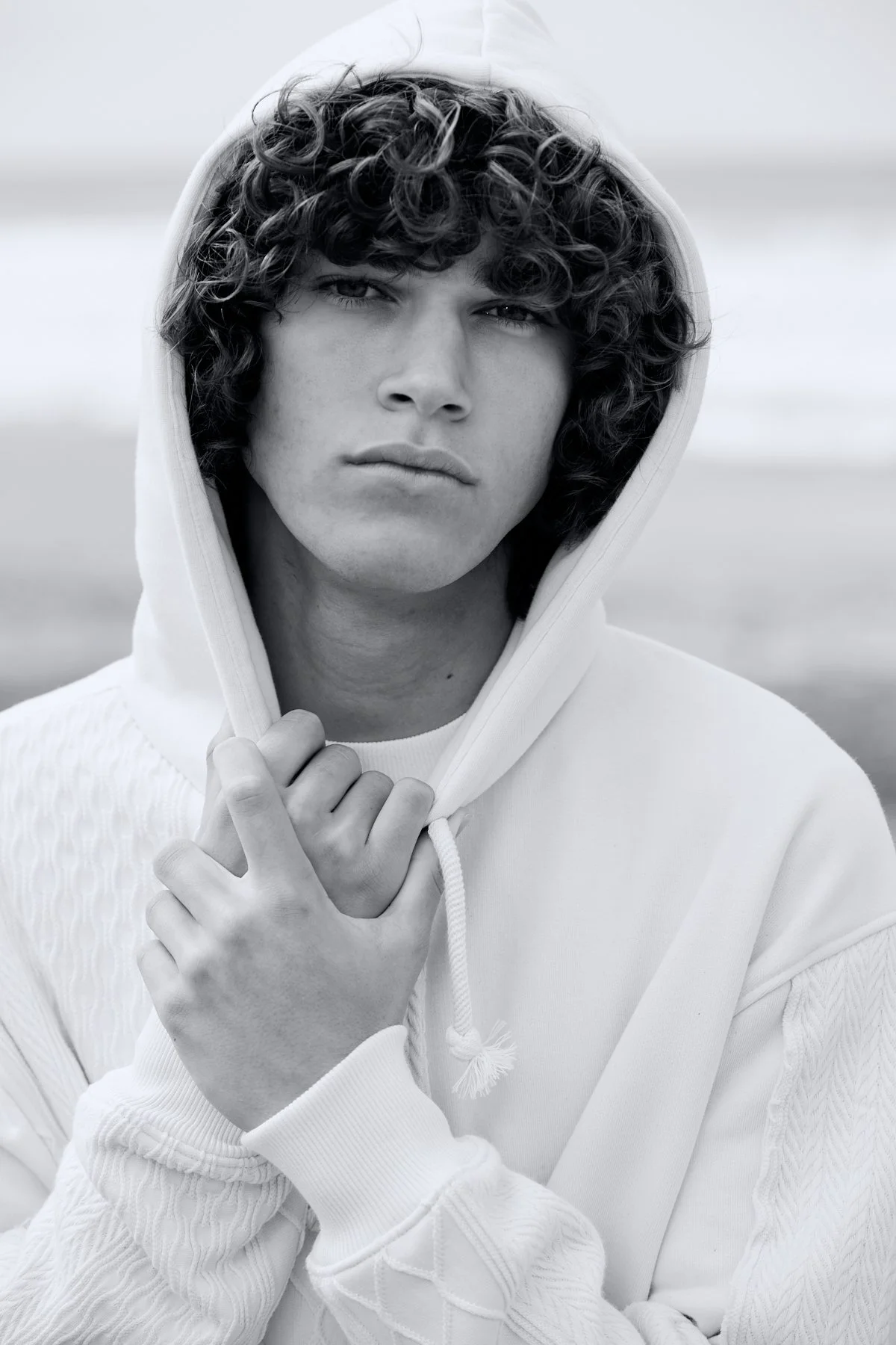 Black and white photo of a young man with curly hair wearing a hoodie, holding the hood near his face, outdoors.