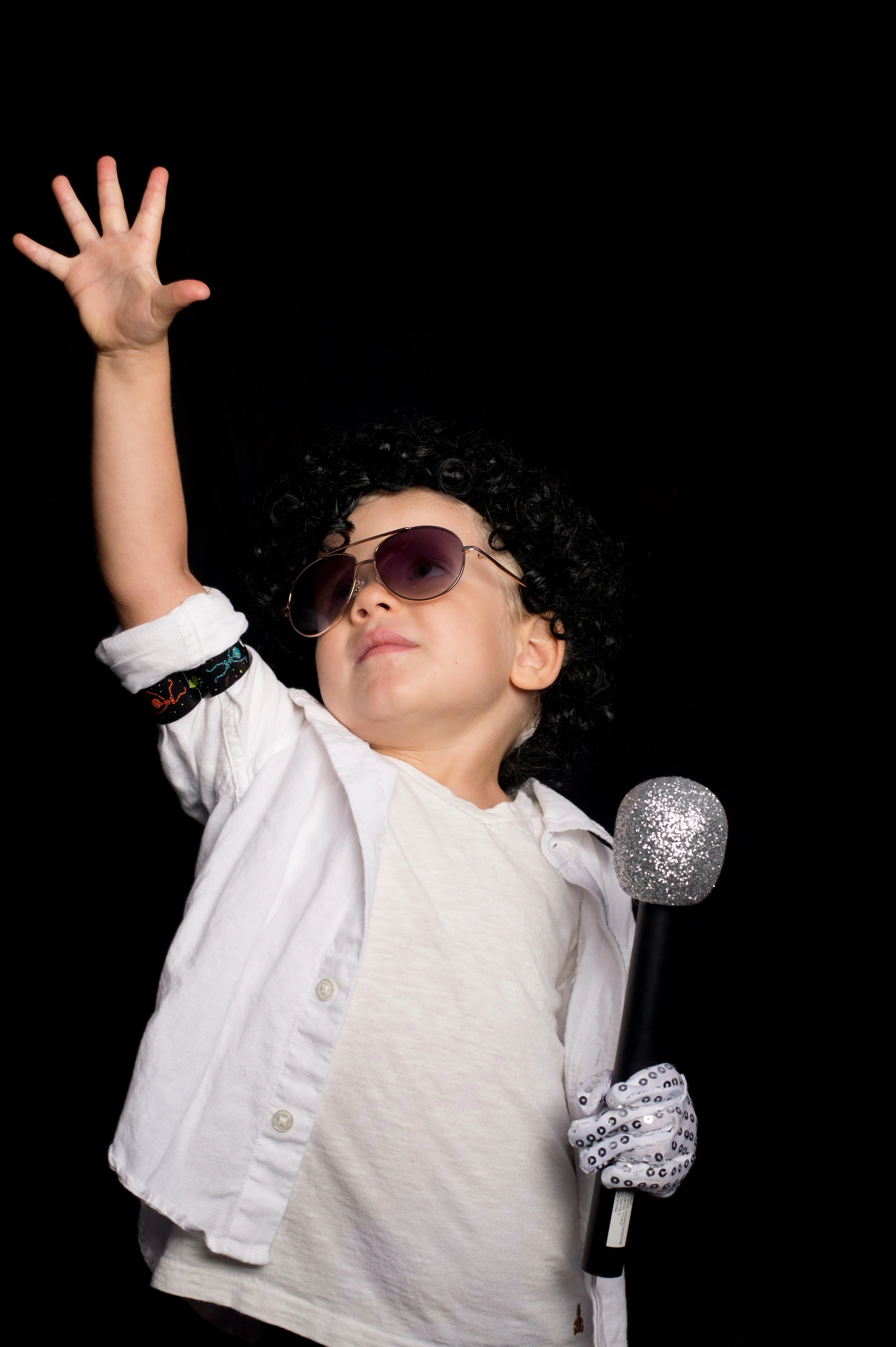 A child with curly hair, wearing sunglasses, a white shirt, and a white jacket, holding a microphone with a glittery silver top, and raising their right hand against a black background.