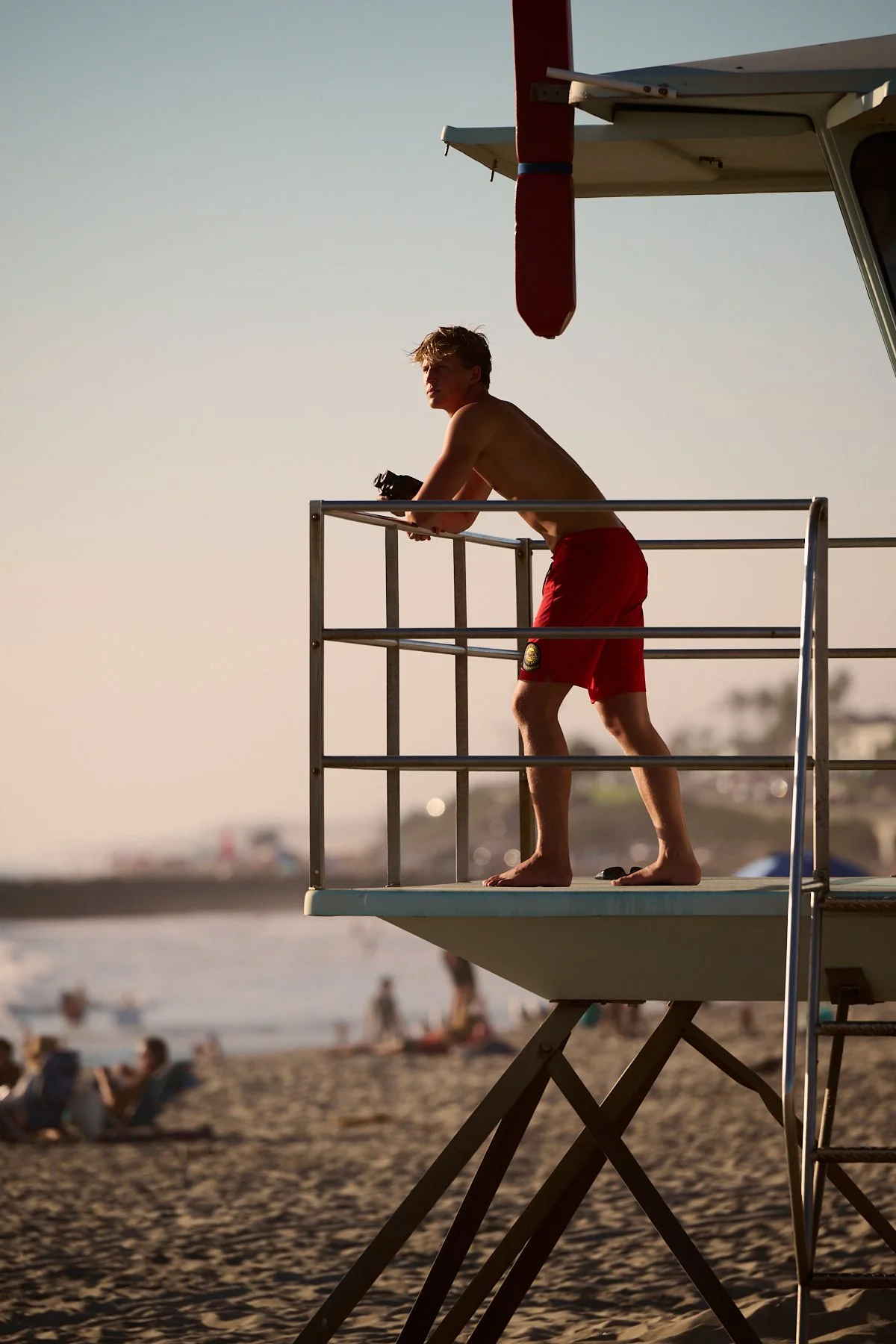 A young man with blonde hair, shirtless and wearing red shorts, standing on a lifeguard tower watching the beach.