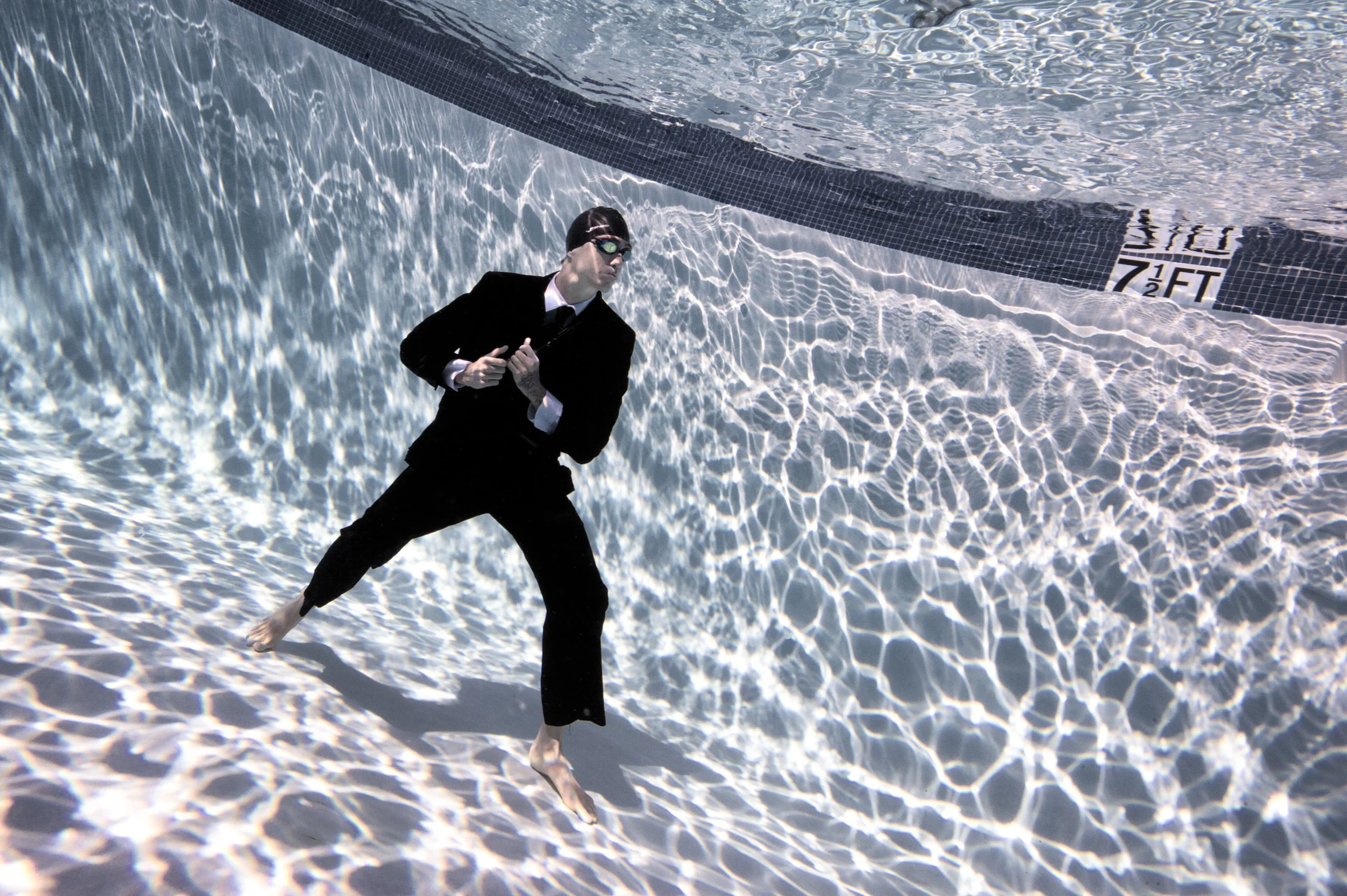 A person in a business suit with goggles stands in a swimming pool, surrounded by water reflections, with the pool's depth marker visible.