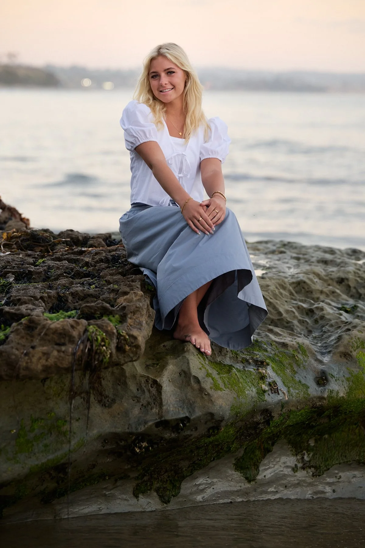 A young woman with blonde hair sitting barefoot on wet rocks near the water at the beach during sunset, smiling at the camera.