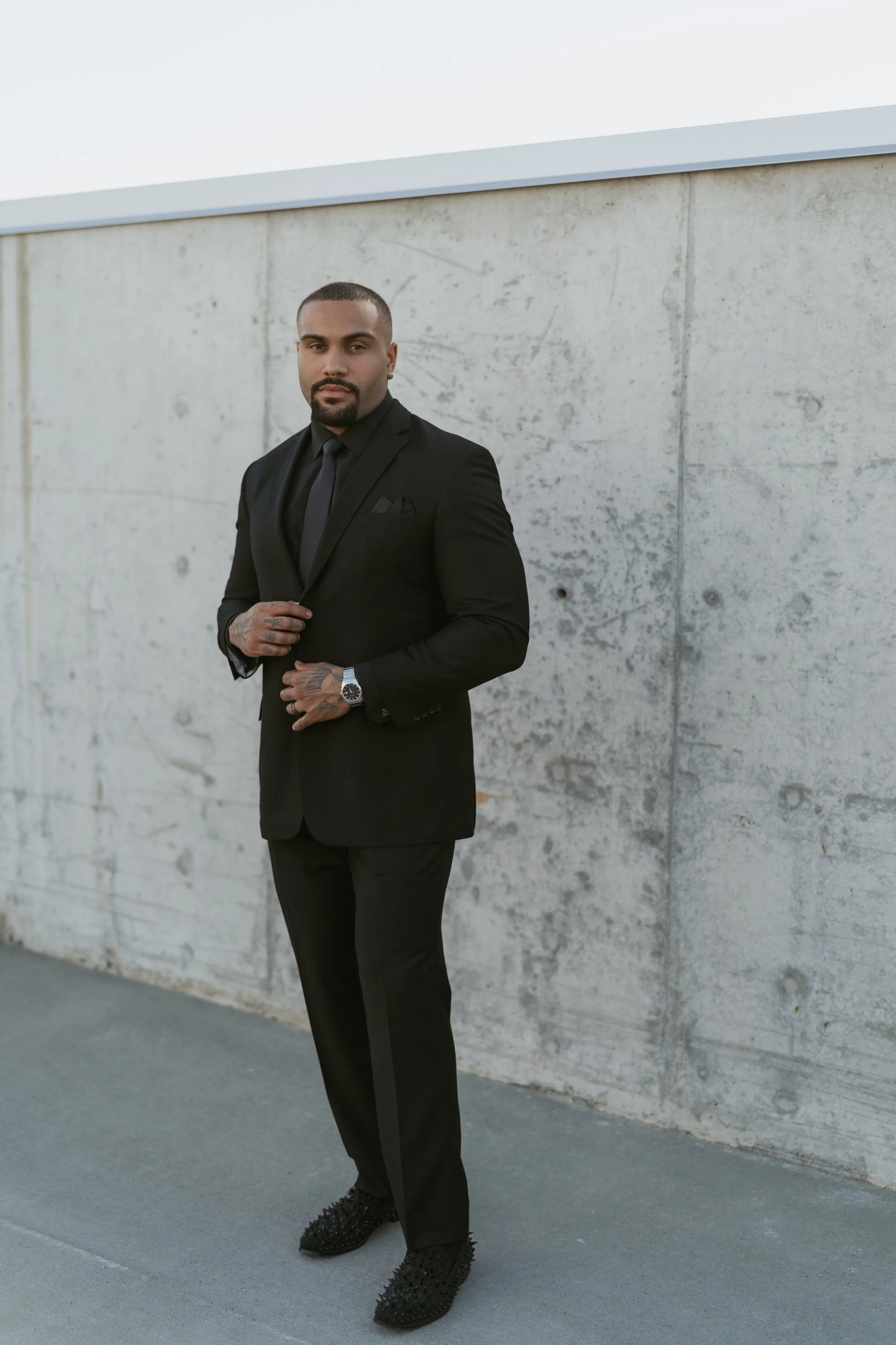 groom in all black suit on wedding day looking directly into the camera very stylish with black shoes  