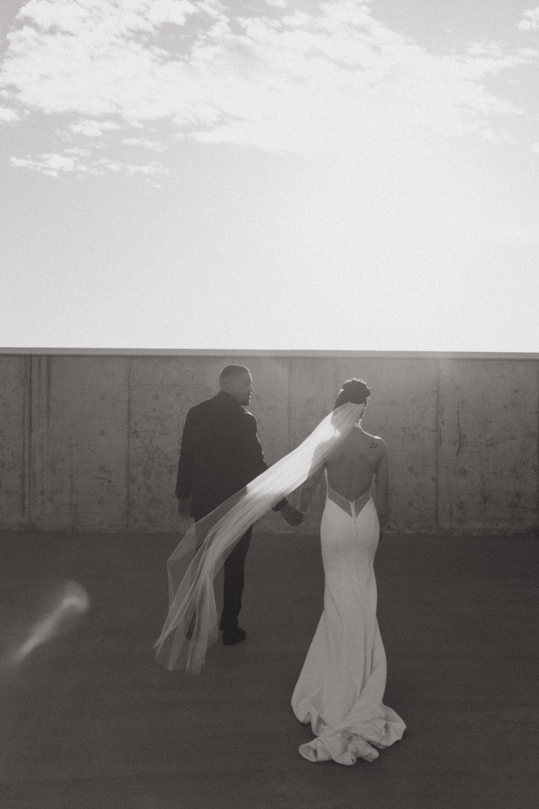 A black-and-white photograph of a bride and groom walking outdoors, with the bride wearing a long gown and veil, and the groom in a dark suit.