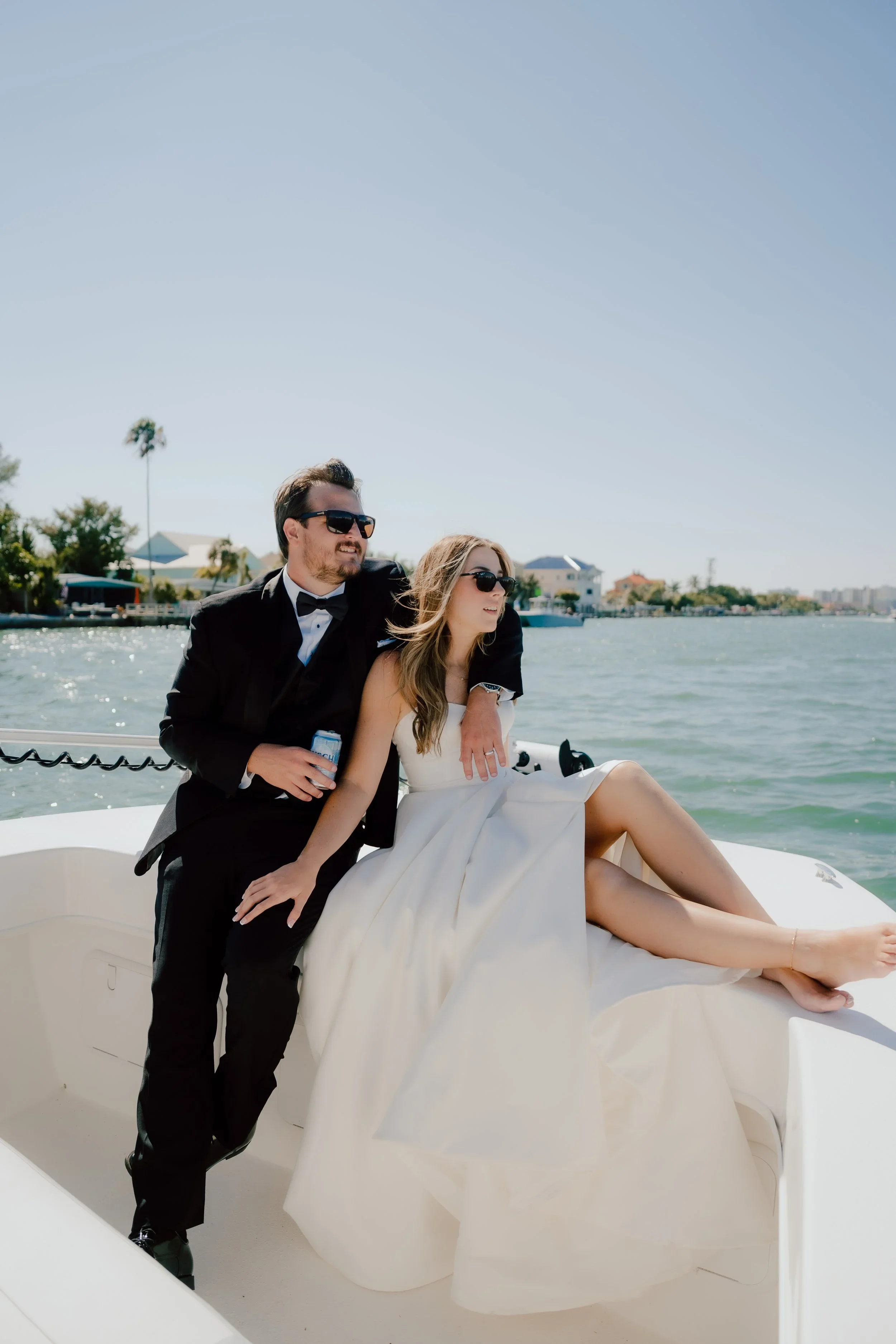 A couple in wedding attire on a boat on a body of water with houses, trees, and clear blue sky in the background.