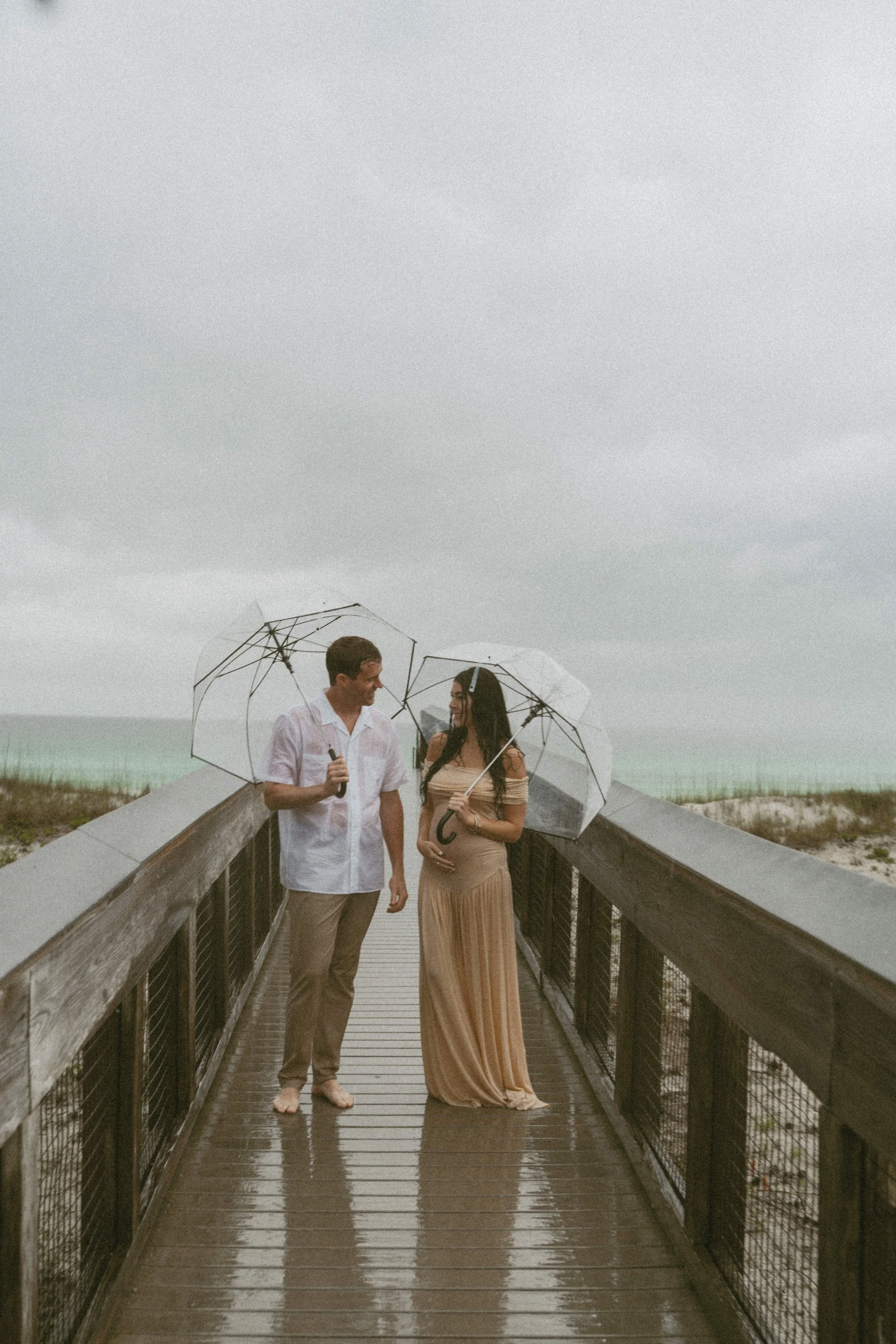 A couple standing on a wooden boardwalk at the beach, holding umbrellas on a rainy day. The man is wearing a white shirt and beige pants, and the woman is in a long beige dress, pregnant, holding her belly. They are smiling and looking at each other.