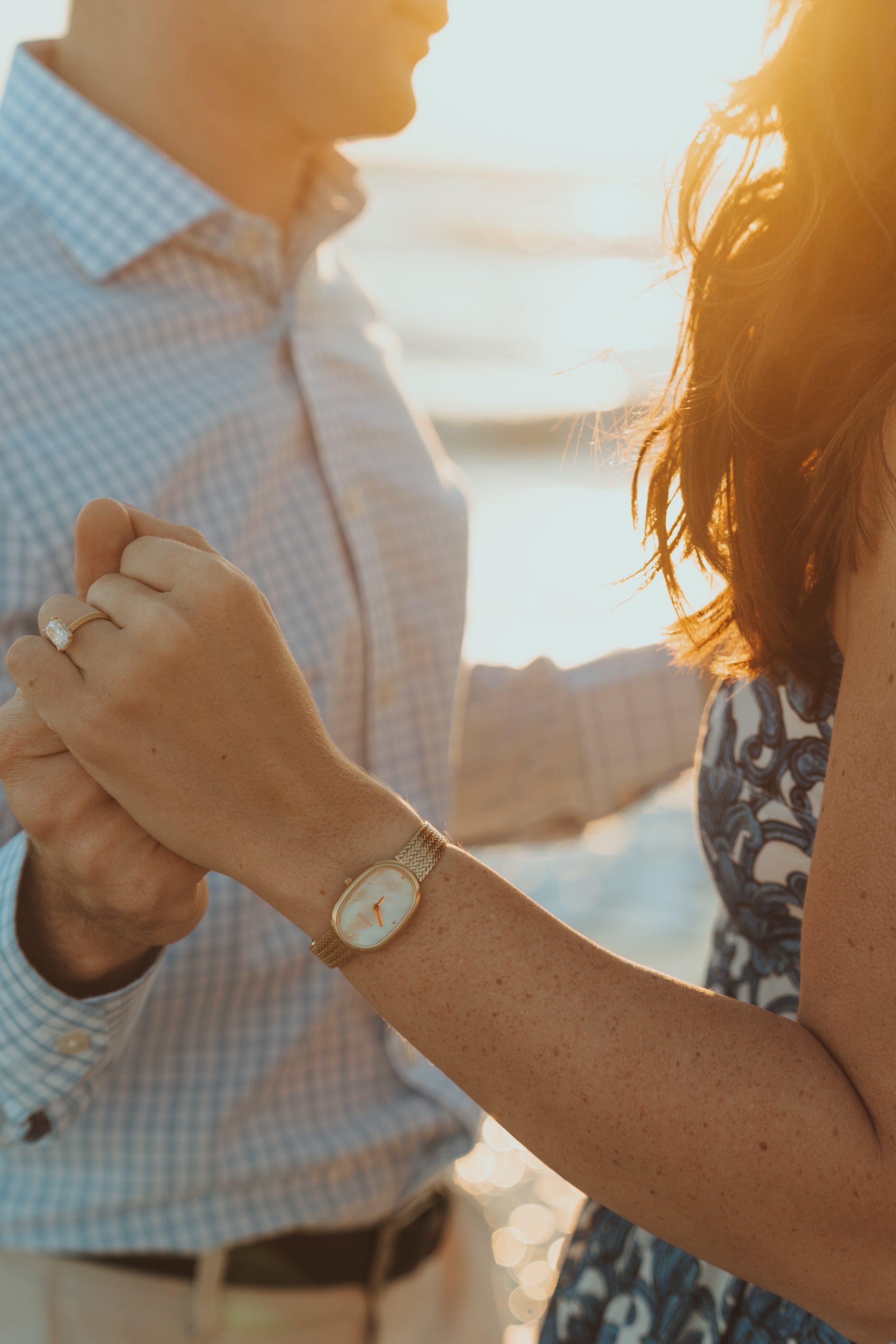A man and woman holding hands during sunset on the Destin Florida beach, with the woman wearing a wristwatch and engagement ring.