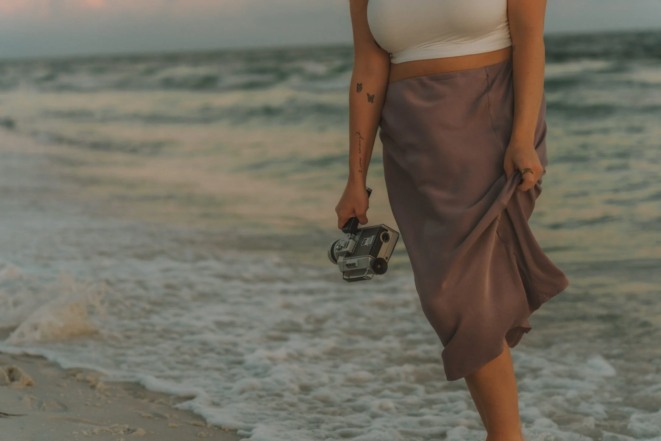 A person walking on the beach holding a vintage camera in their right hand, wearing a white crop top and a long brown skirt, with visible tattoos on their forearm.