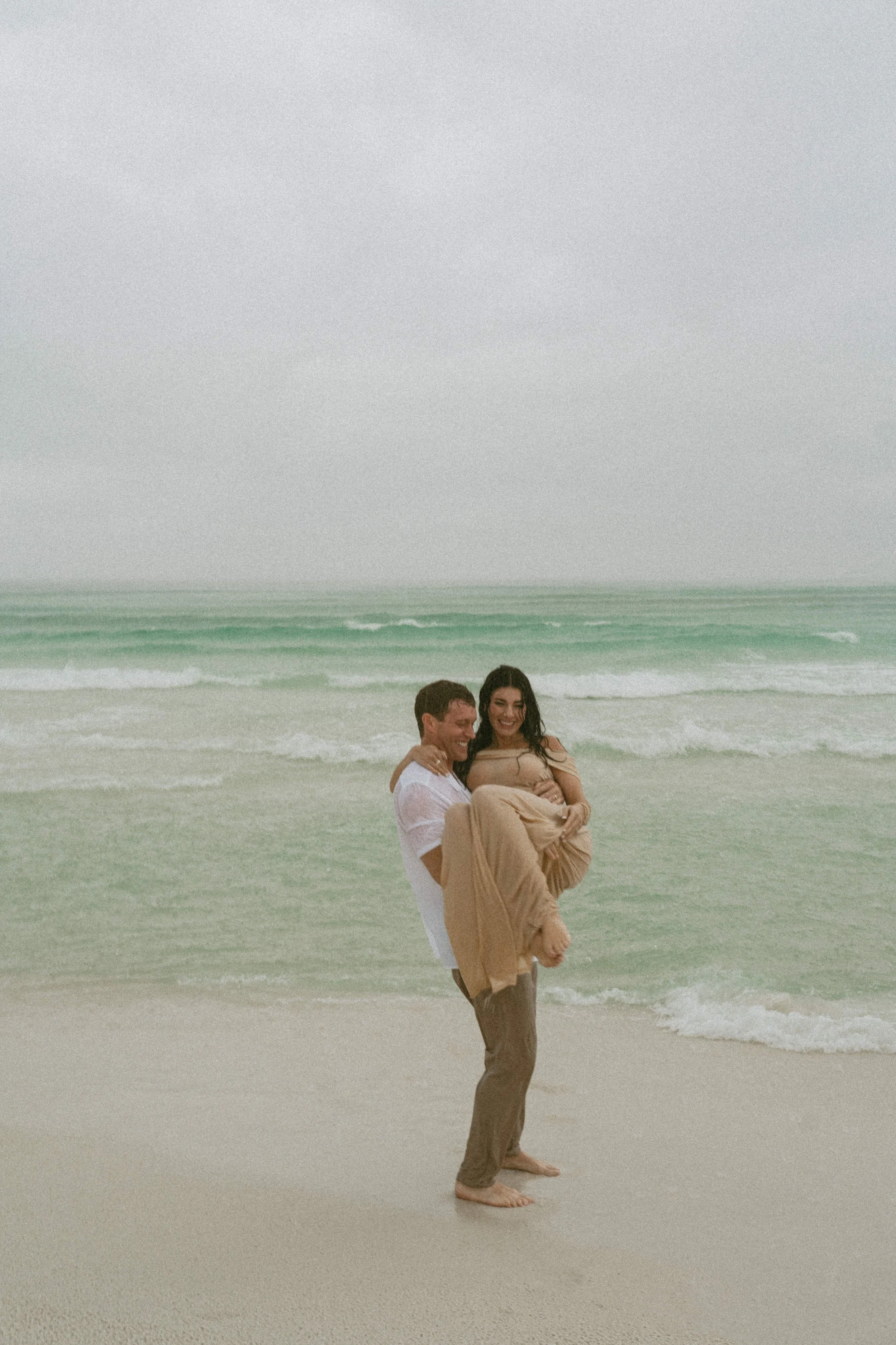 A man holding a woman in his arms at the beach with waves in the background.