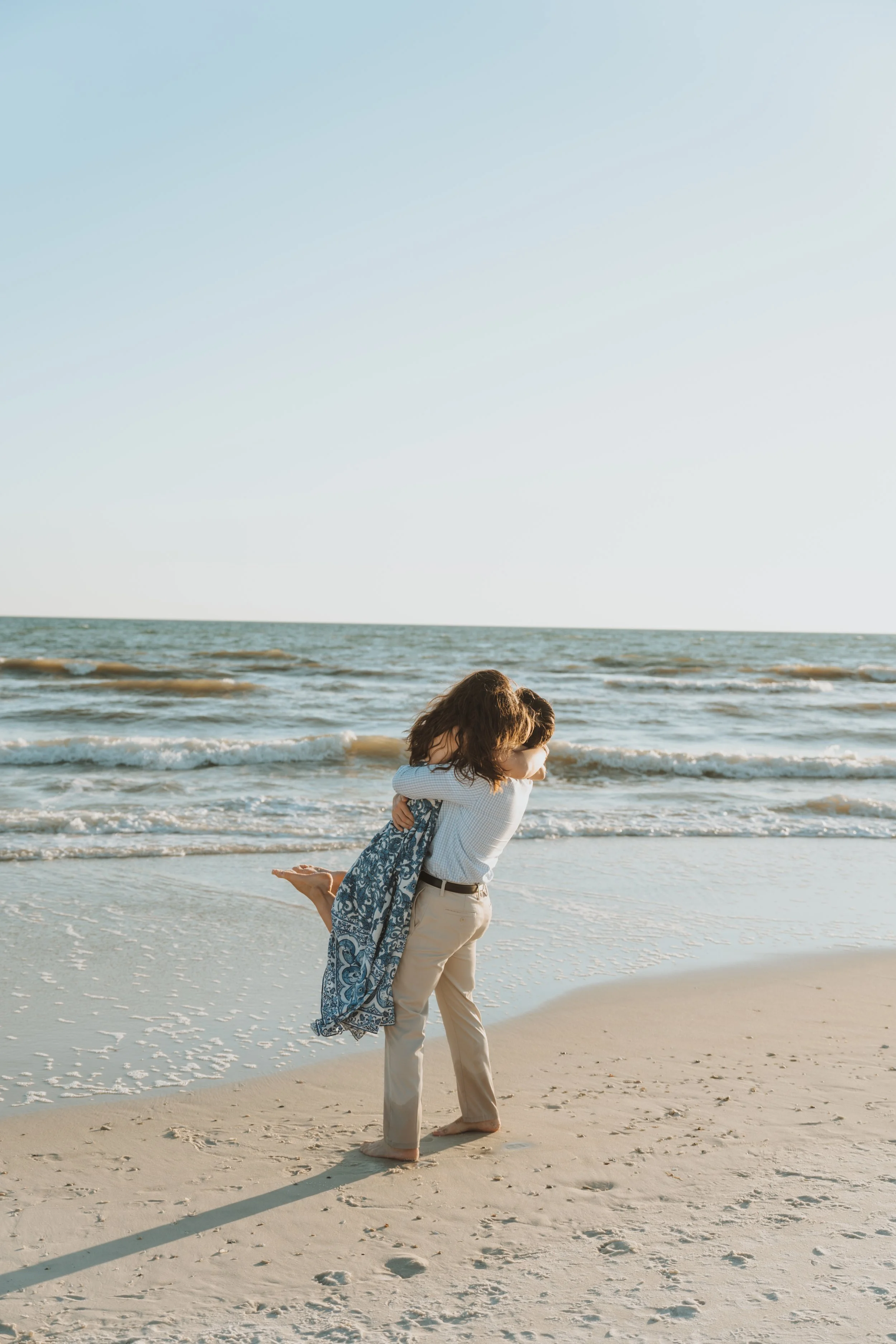 A couple hugging on a sandy beach with waves and a clear sky in the background.