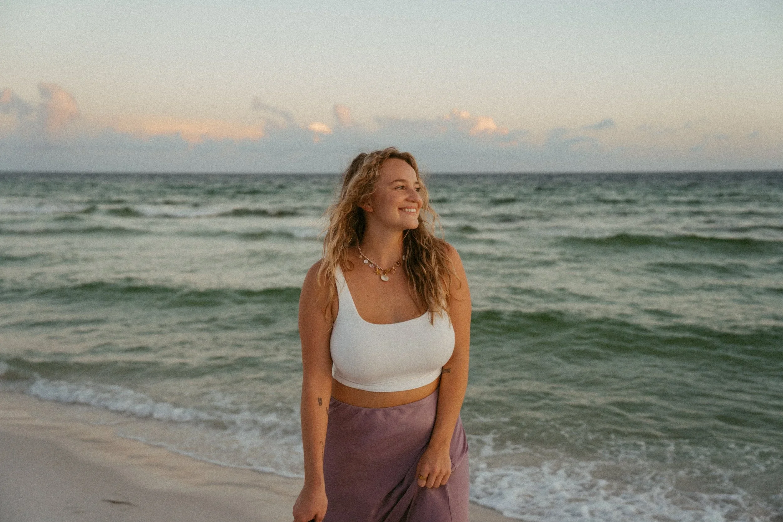 A woman with curly blonde hair stands on the beach near the water, smiling and looking to the side, wearing a white tank top, purple skirt, and layered necklaces.