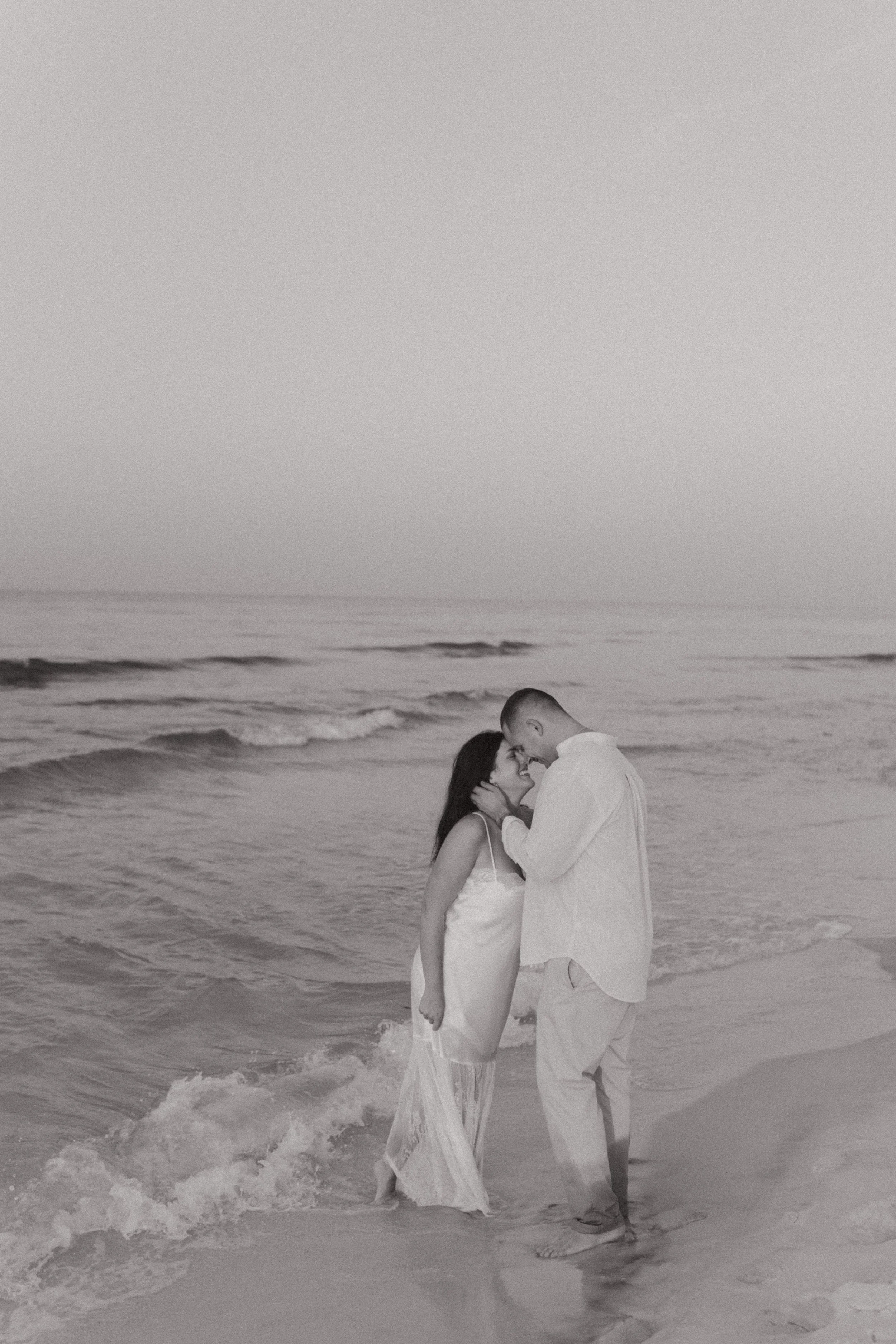 A couple dressed in white clothes sharing an intimate moment with foreheads touching on the beach during sunset, with gentle waves in the background.
