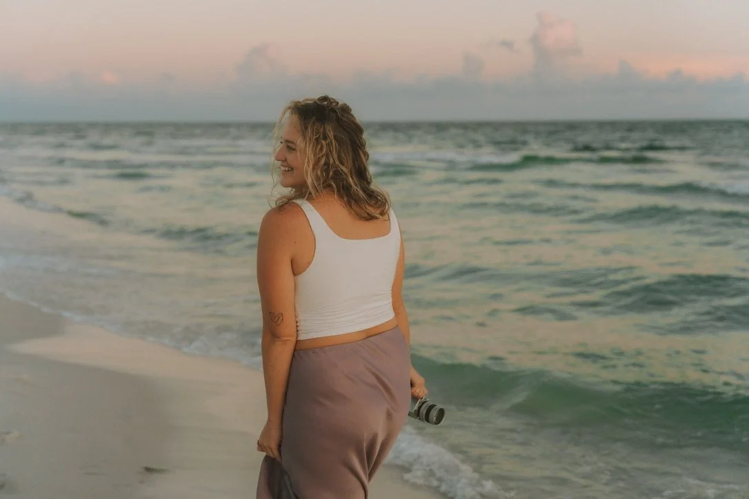 A woman with curly hair smiling on a beach during sunset, holding a camera, wearing a white tank top and beige pants.