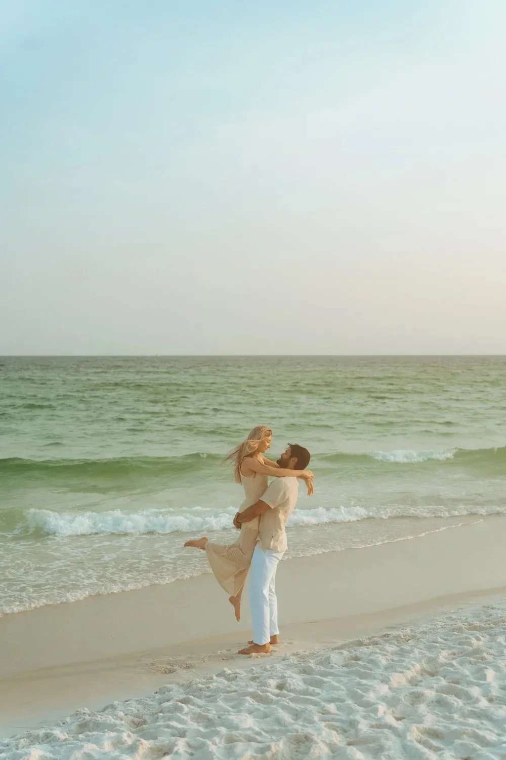 A couple on the beach with the man lifting the woman in his arms near the shoreline, ocean waves in the background, and a clear sky.