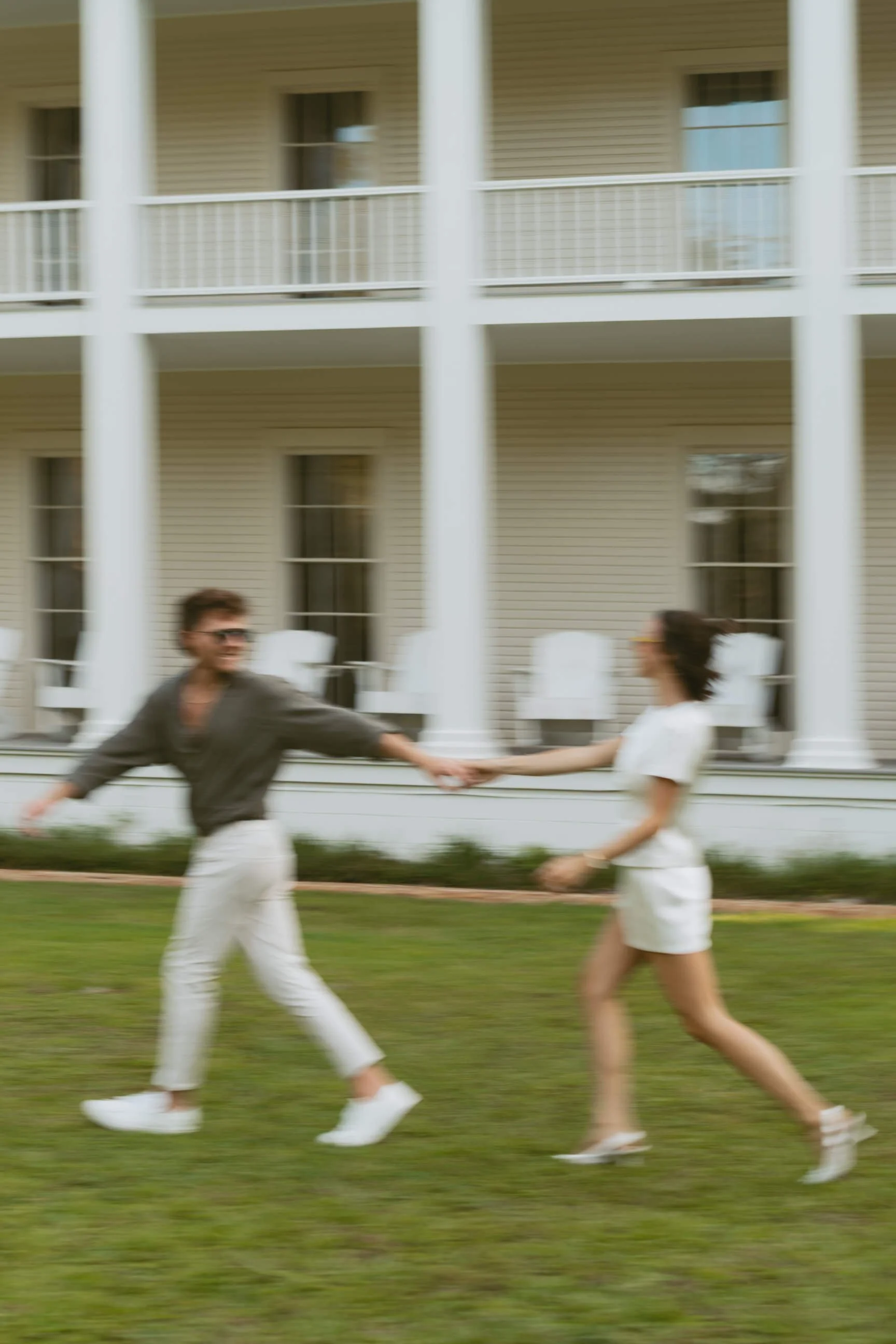 A man and woman holding hands and playing on the grass in front of a large house with white columns and railings, with the scene blurred due to motion.