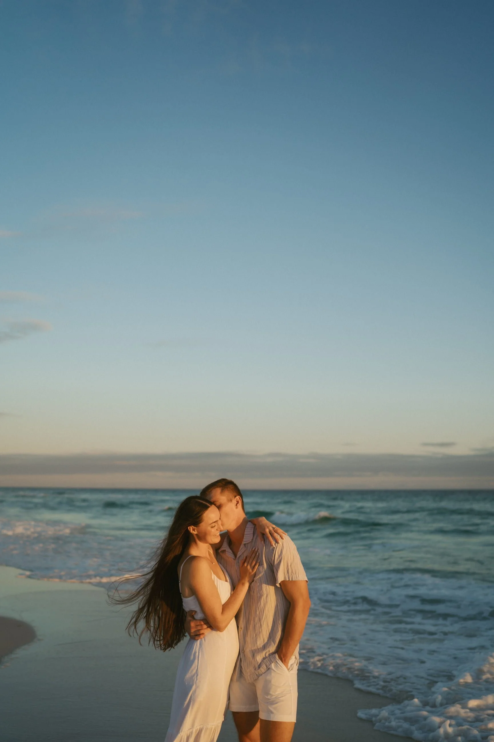 A couple embracing and kissing on the beach during sunset with ocean waves in the background.