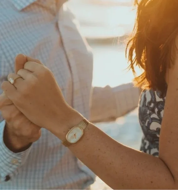 Two people holding hands on a beach at sunset, with one person wearing a gold watch and a ring.