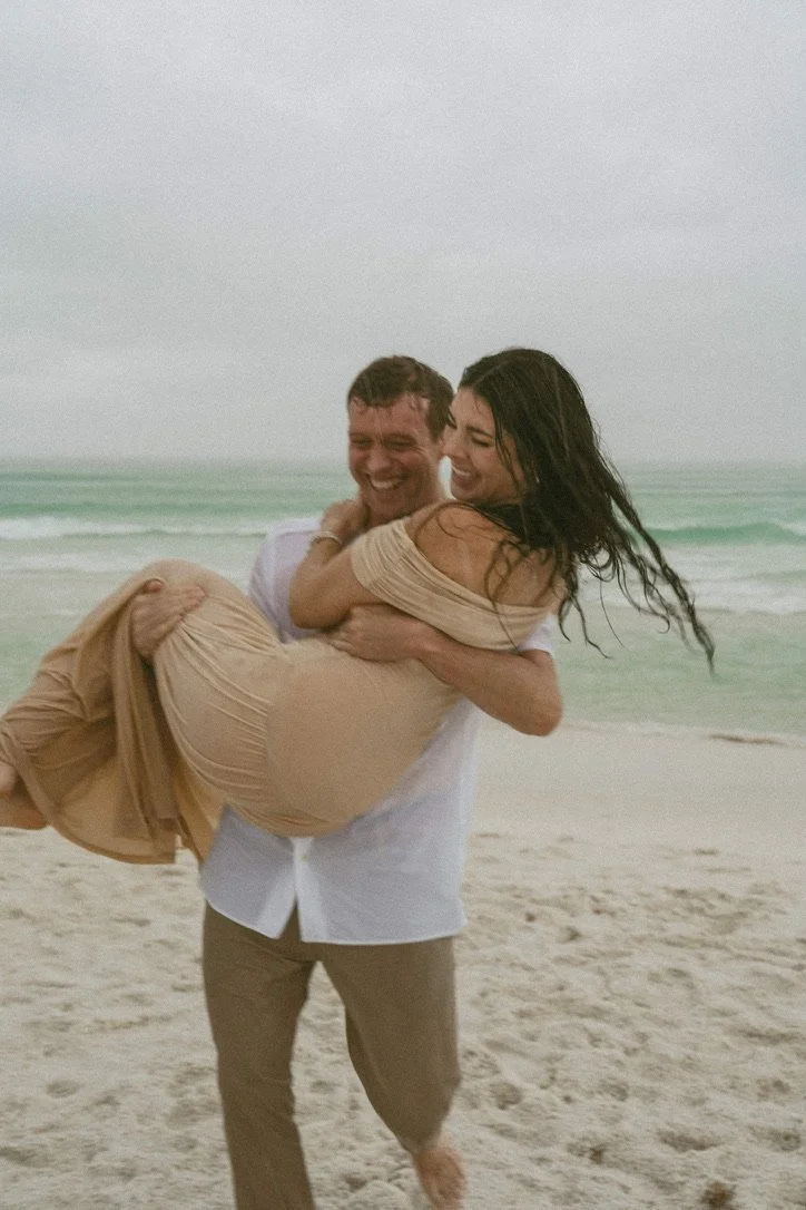 A couple is on the beach with the ocean in the background. The man is lifting the woman in his arms, and they are smiling and laughing.