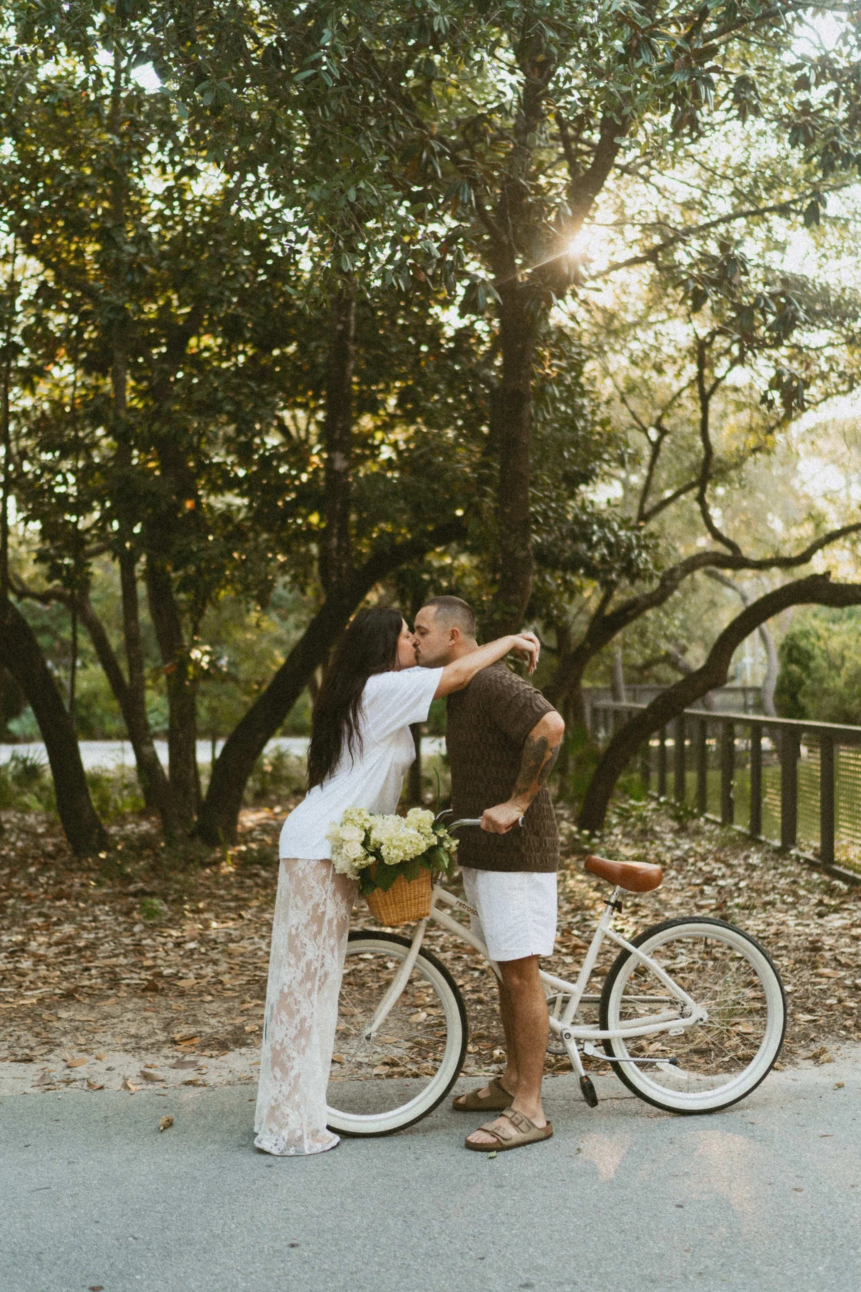 A couple kissing outdoors beside a bicycle with a basket of white flowers, in a park-like setting with trees and sunlight.