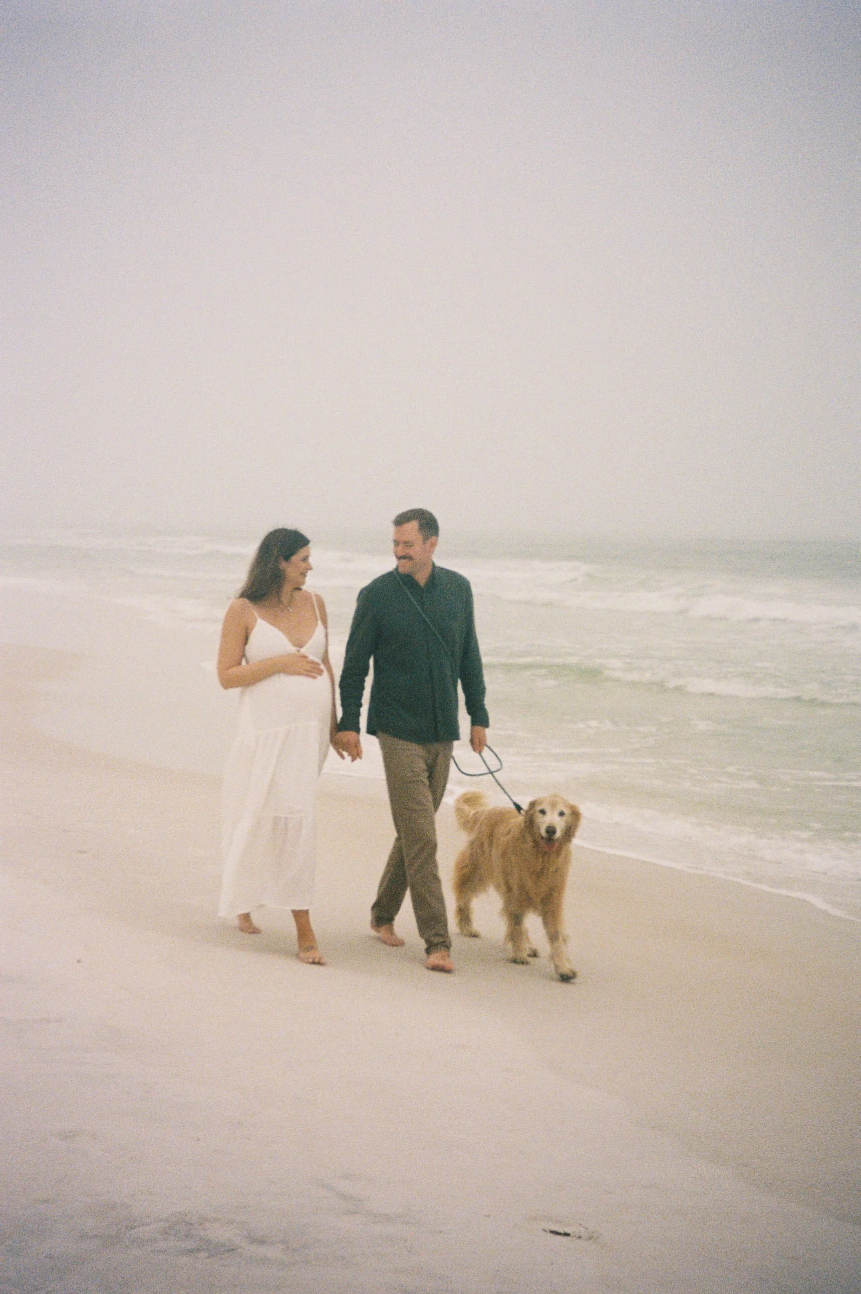 A couple walking along the beach with their dog, holding hands and smiling.