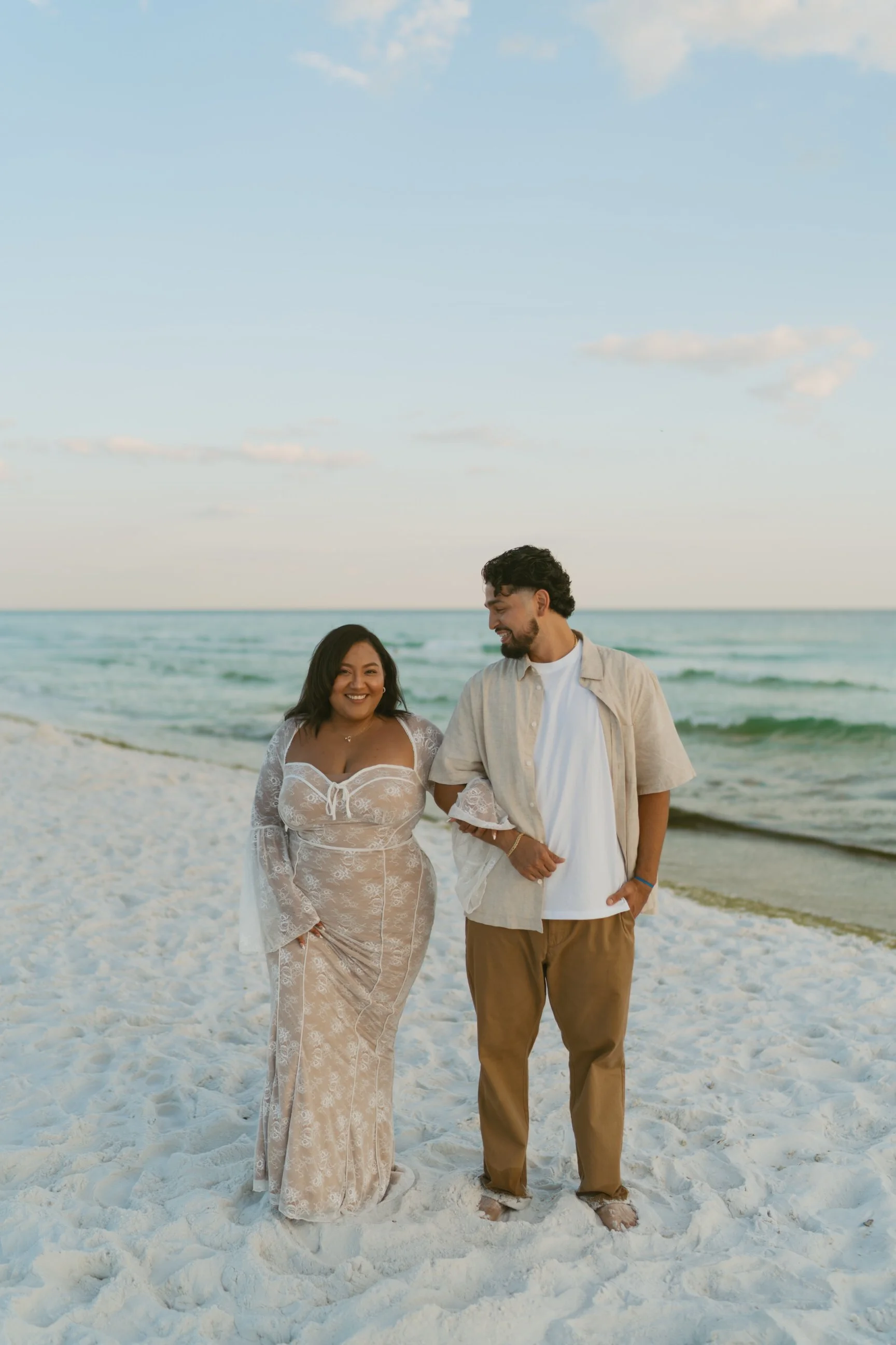 A couple walking on the beach, smiling, with the ocean and sky in the background.