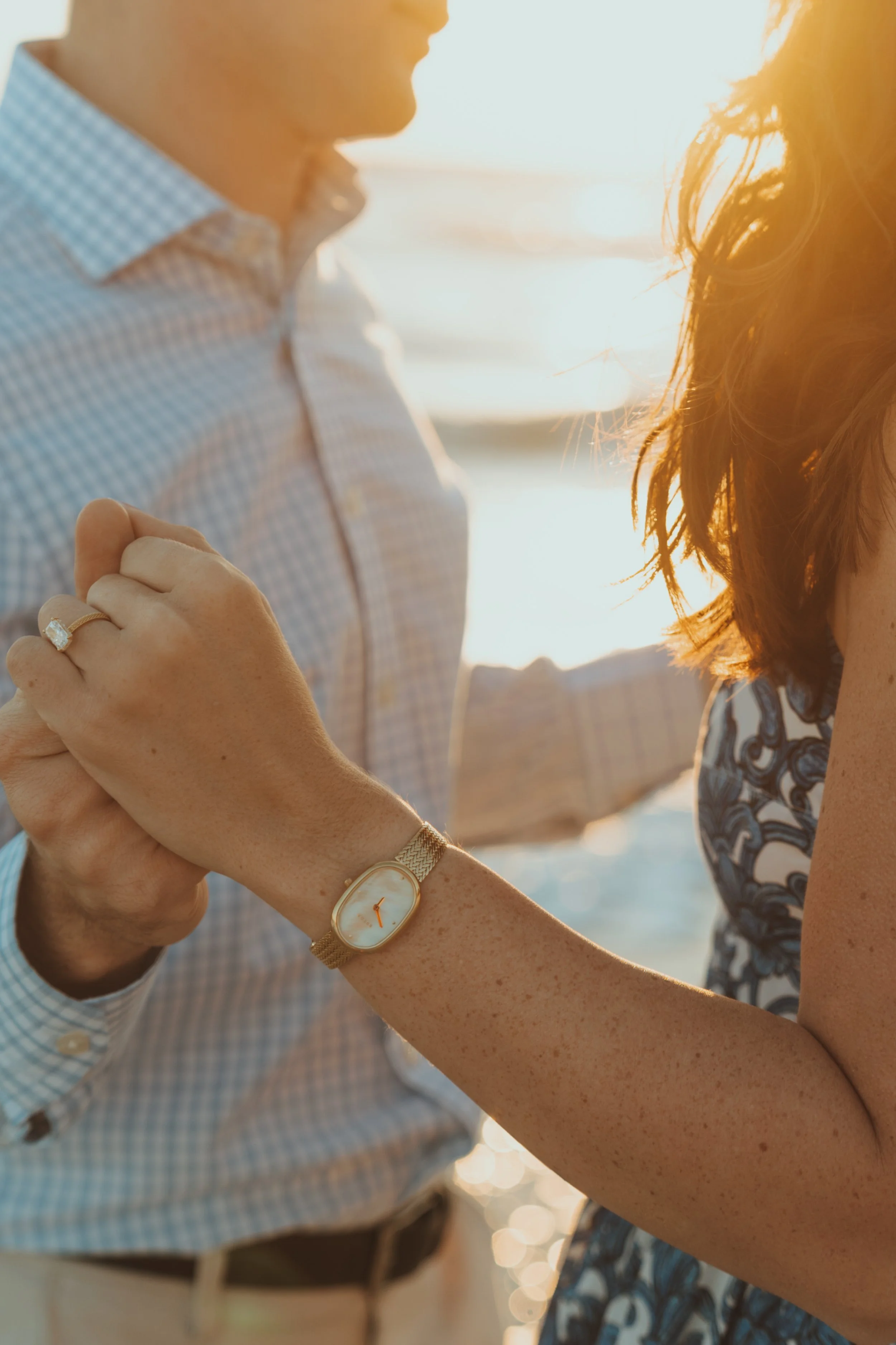 A man and woman holding hands by the water during sunset, with the woman wearing a gold watch and ring.