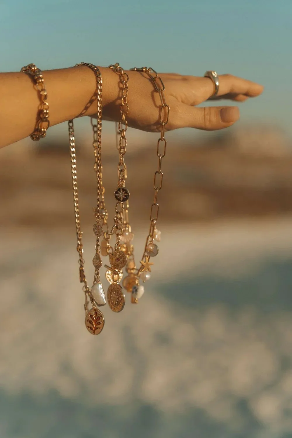 Hand holding several gold chains and necklaces with various charms against a beach background.