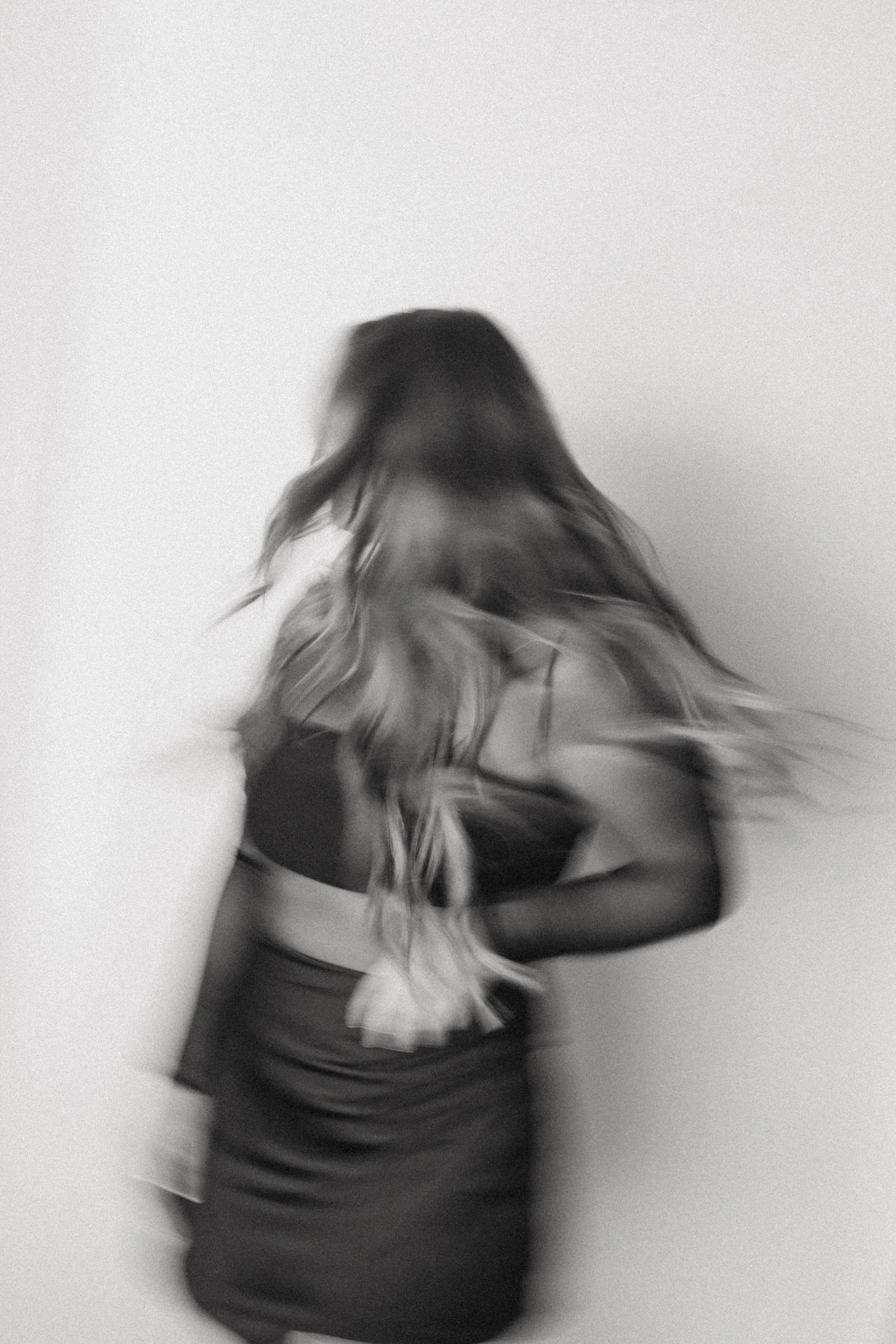 A blurred black and white photo of a woman with long, wavy hair wearing a dress and black gloves, standing against a plain light-colored wall.
