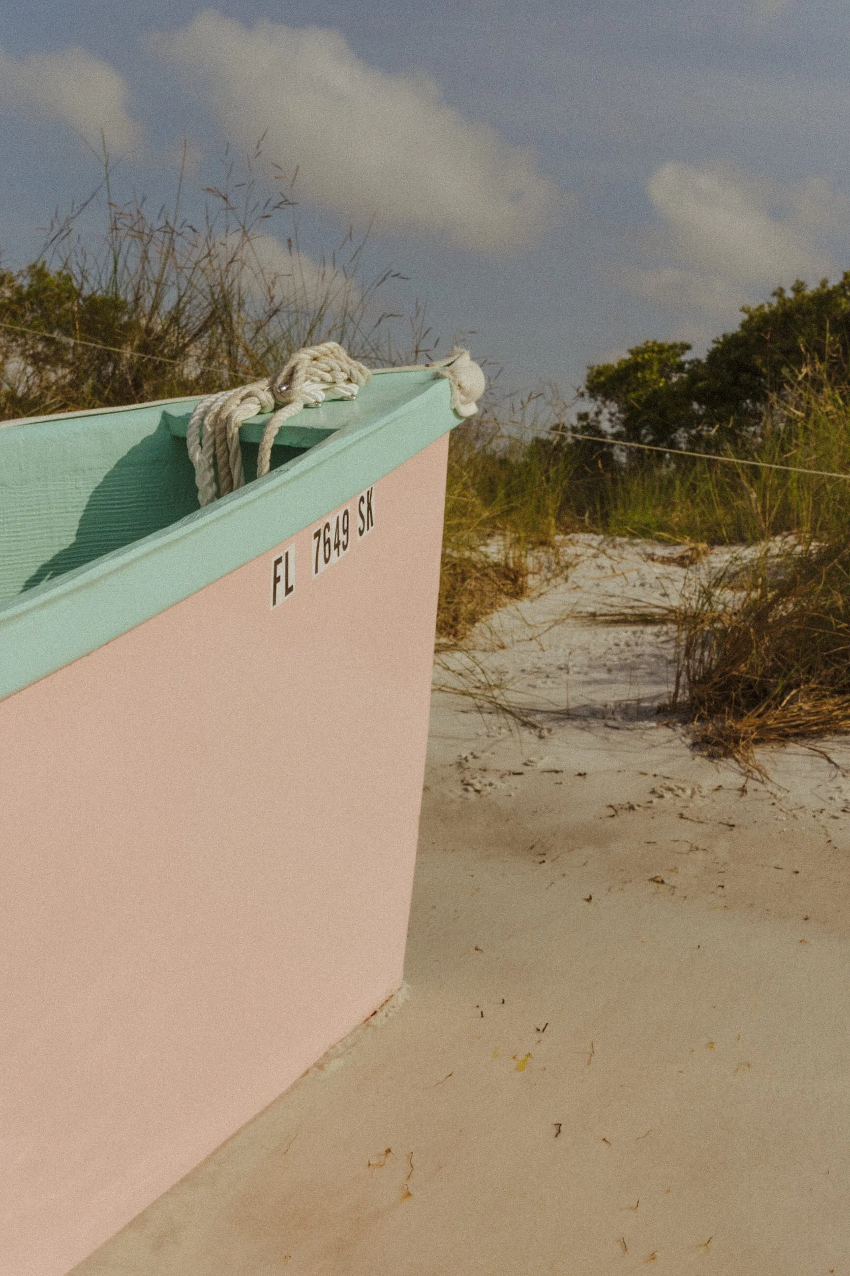 Close-up of a pastel pink and mint green boat on a sandy beach with beach vegetation and cloudy sky in the background.