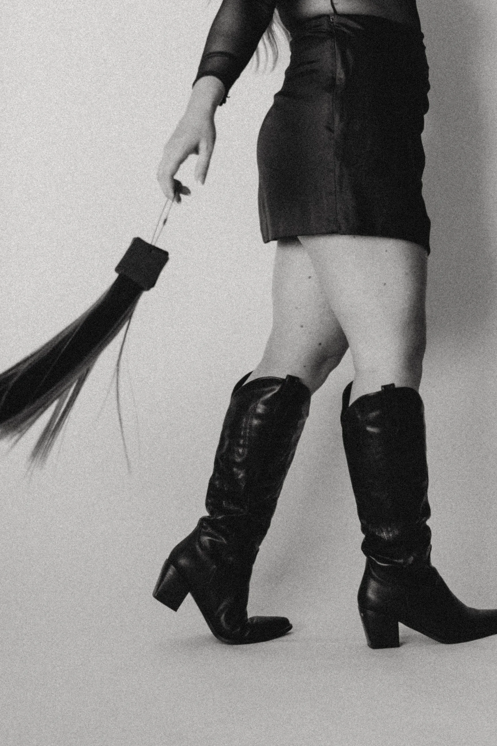 A woman wearing a black top, short black skirt, and tall black leather boots stands against a plain wall, holding hair extensions
