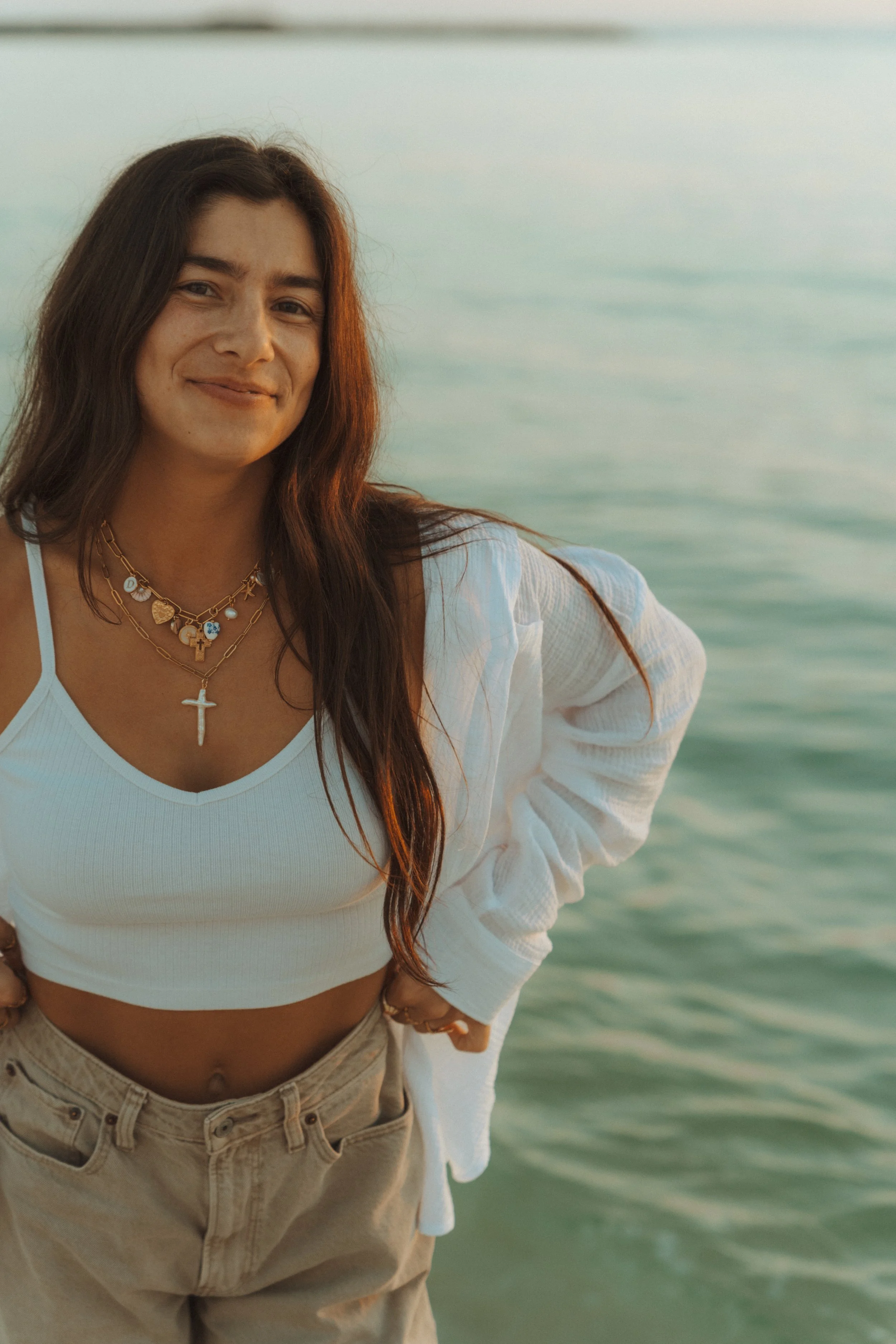 Woman with long dark hair wearing layered necklaces, a white crop top, and beige pants, standing by water with a calm expression.