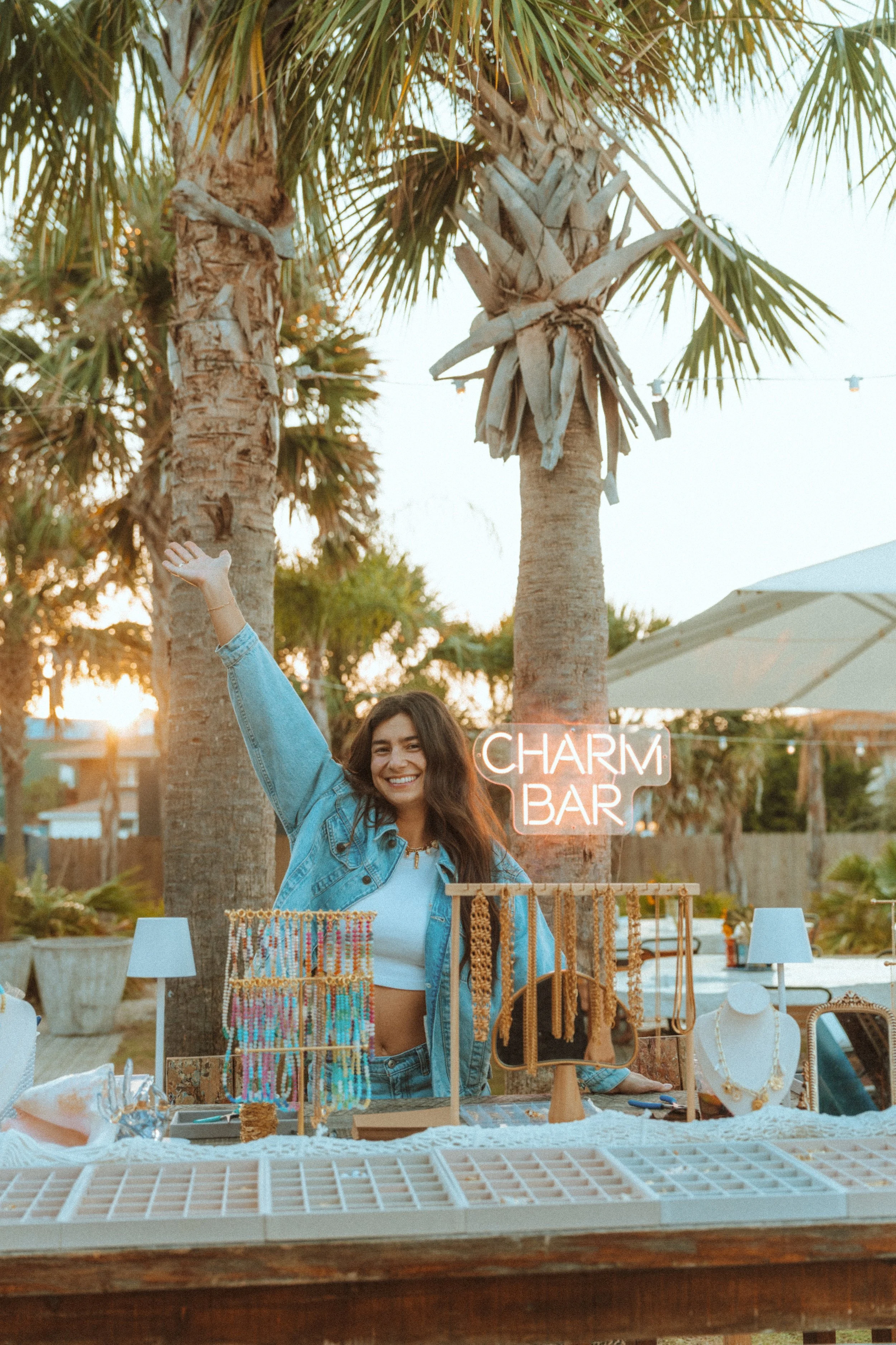A smiling young woman with long dark hair, wearing a denim jacket over a white top, standing at a jewelry stall with a neon sign that reads 'CHARM BAR' in a tropical outdoor setting with palm trees and a sunset.
