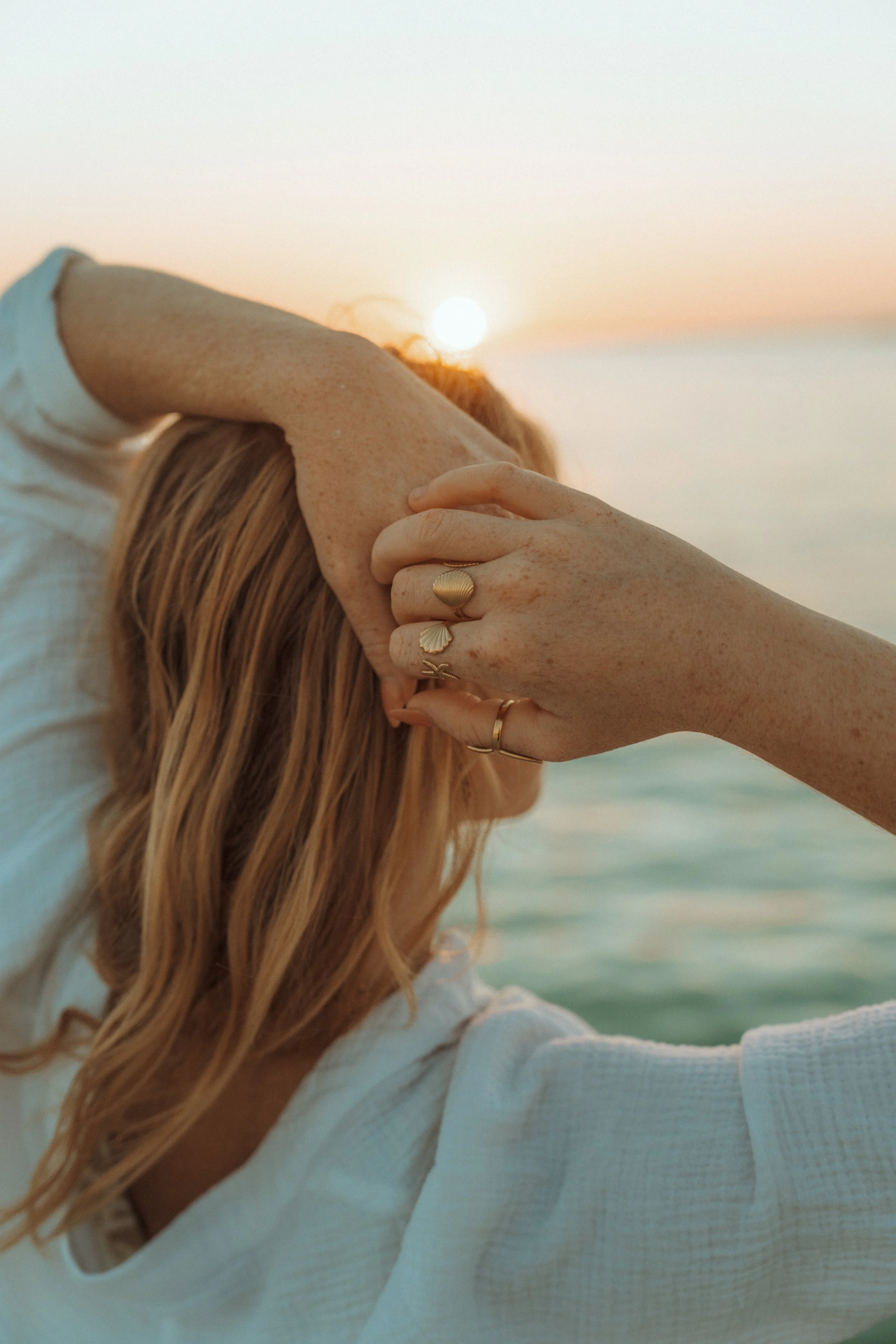A person with freckled skin and red hair is at the beach during sunset, wearing a white top, with one arm resting on their head and the other hand covering their face. They are wearing multiple rings, including a shell-shaped ring.