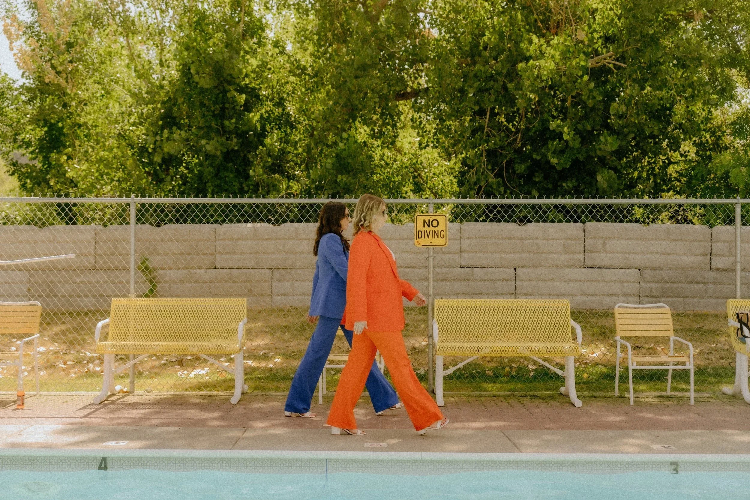 Two women walking along a poolside, one dressed in a blue suit and the other in an orange suit, passing a 'No Diving' sign with green trees and a fence in the background.