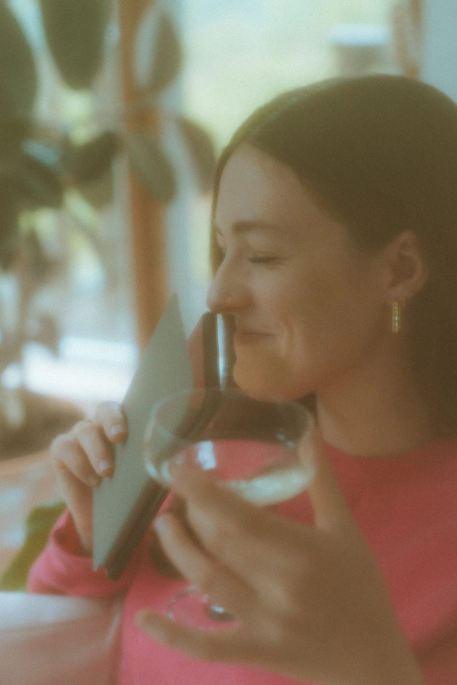A woman with dark hair, wearing a pink top and gold earrings, smiling with her eyes closed, holding a glass of red wine and a mobile phone to her ear, sitting indoors near a window with trees and greenery visible outside.