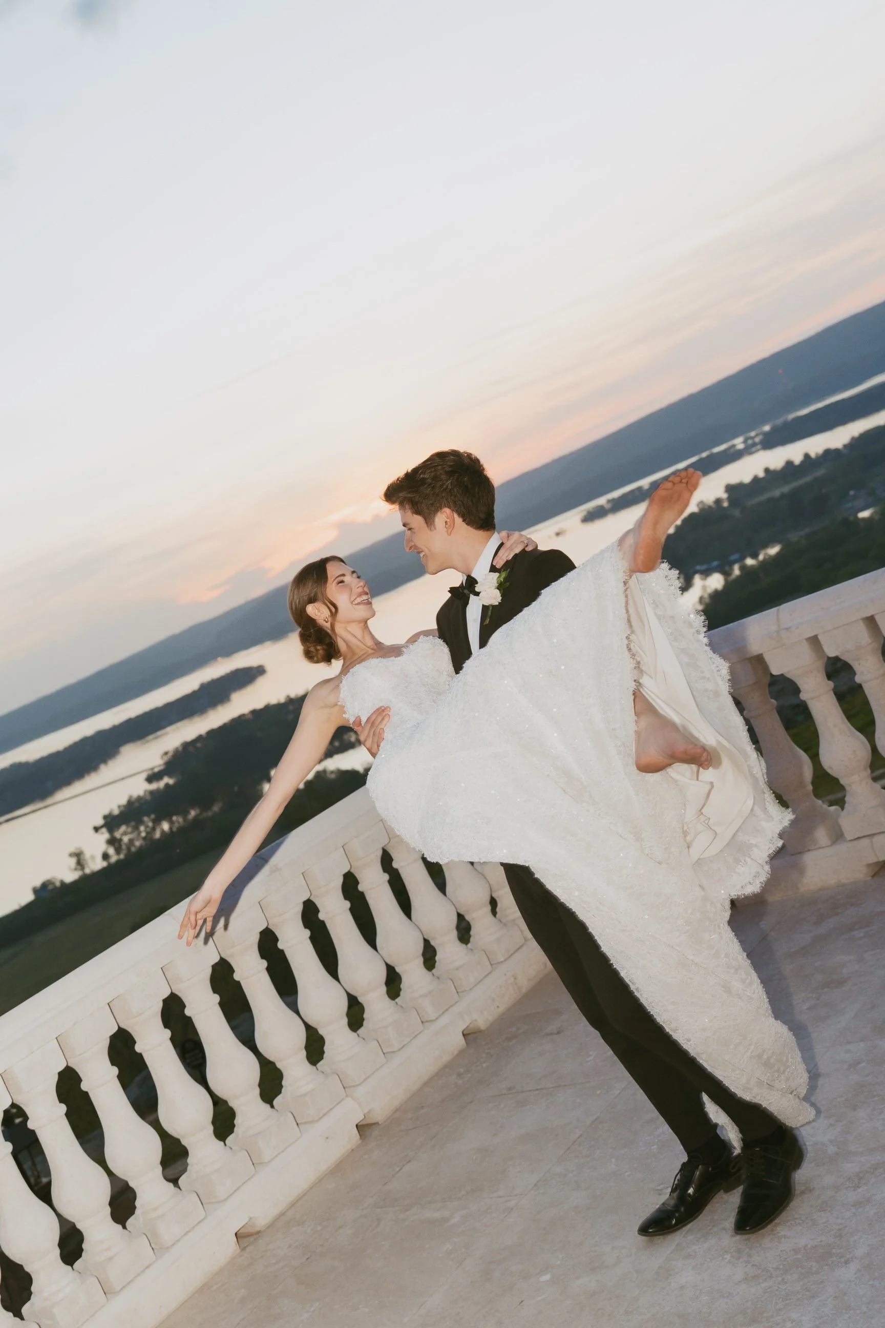 A bride and groom dancing on a balcony during sunset, with a lake in the background.