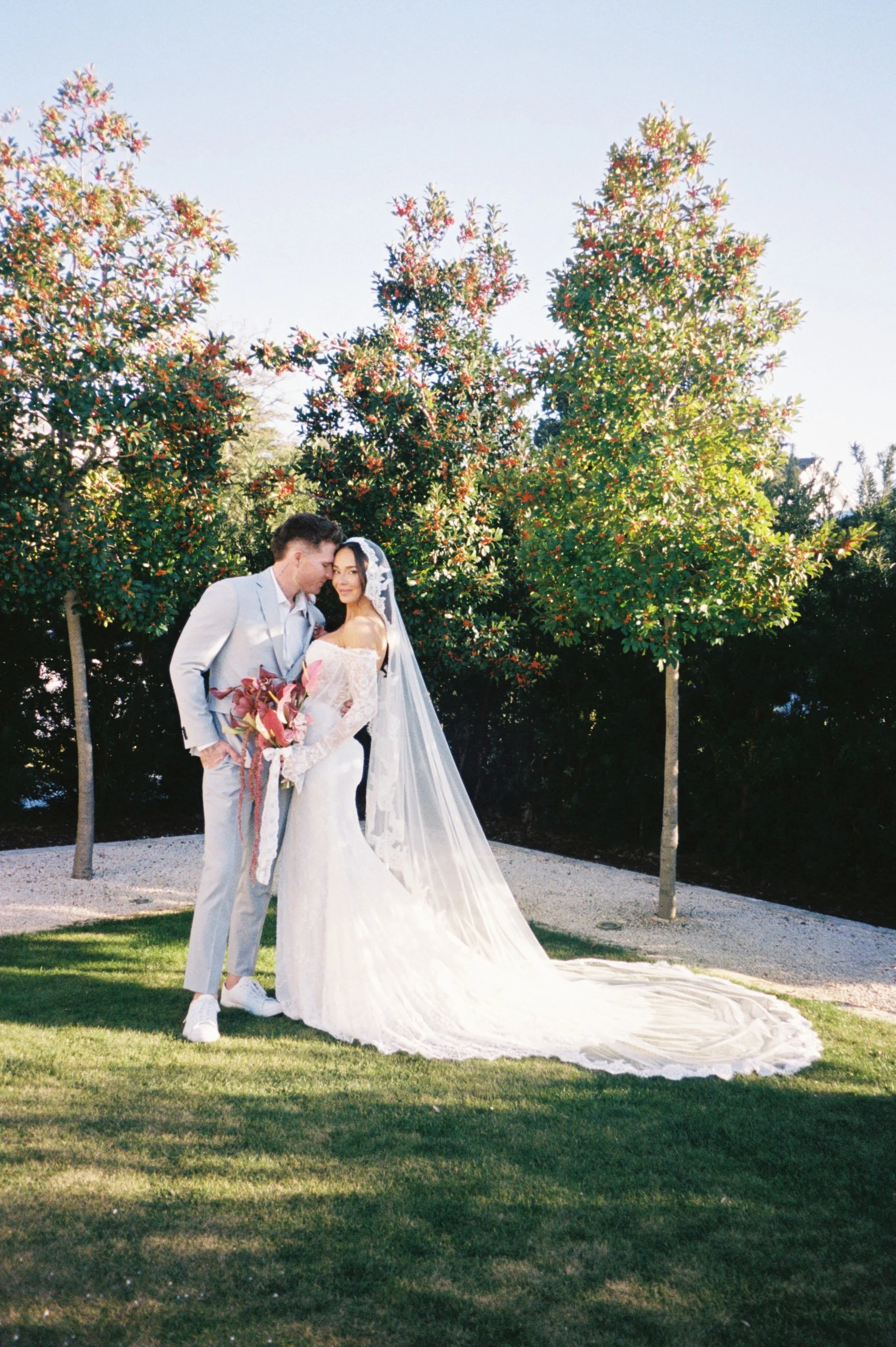 A couple in wedding attire standing outdoors on grass with trees in the background, embracing each other; the bride holds a bouquet, and the groom is leaning his head toward her.