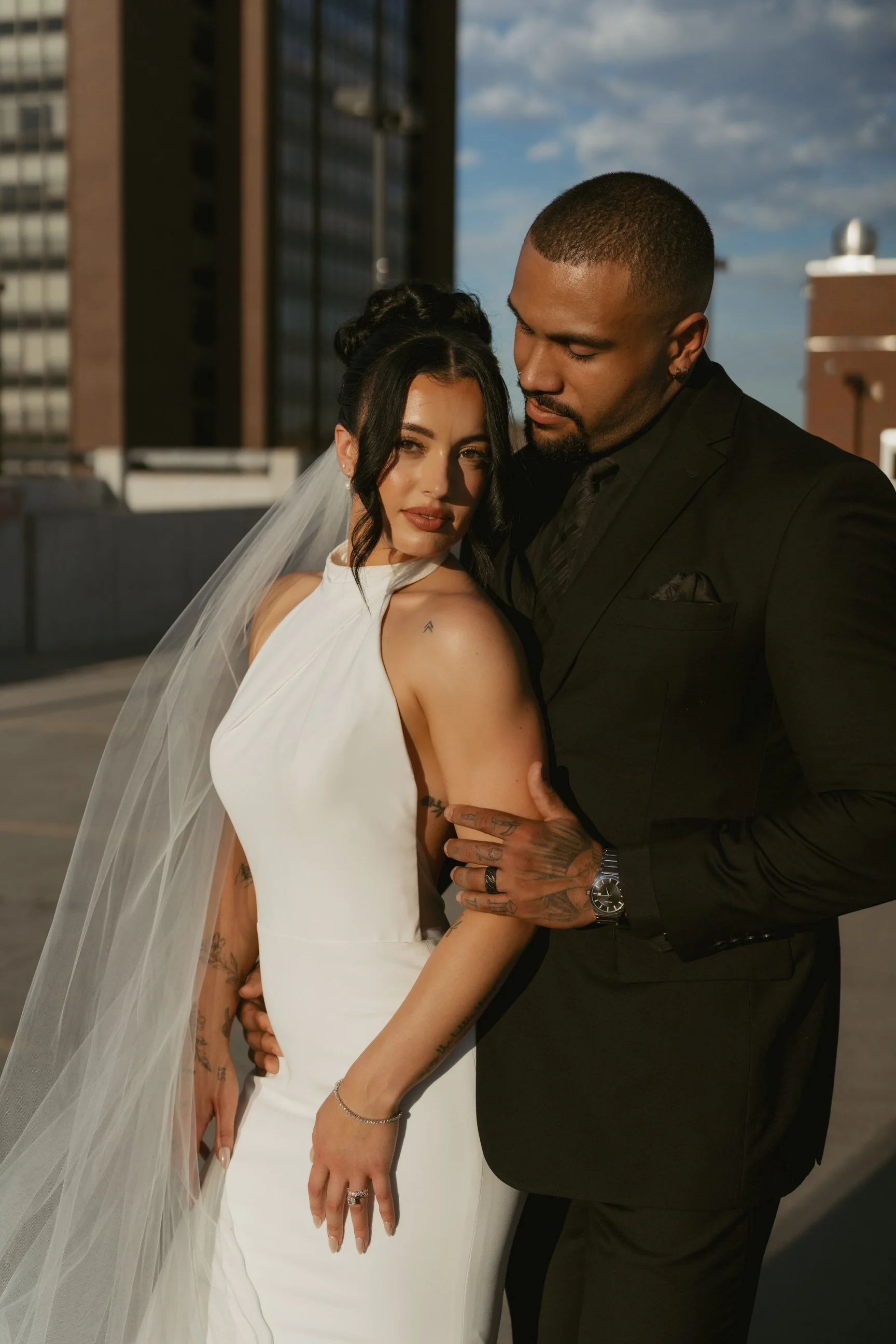 A newlywed couple in wedding attire, standing close together on a rooftop, with the woman wearing a white wedding dress with a veil and the man in a black suit, both looking at the camera with an urban cityscape background.