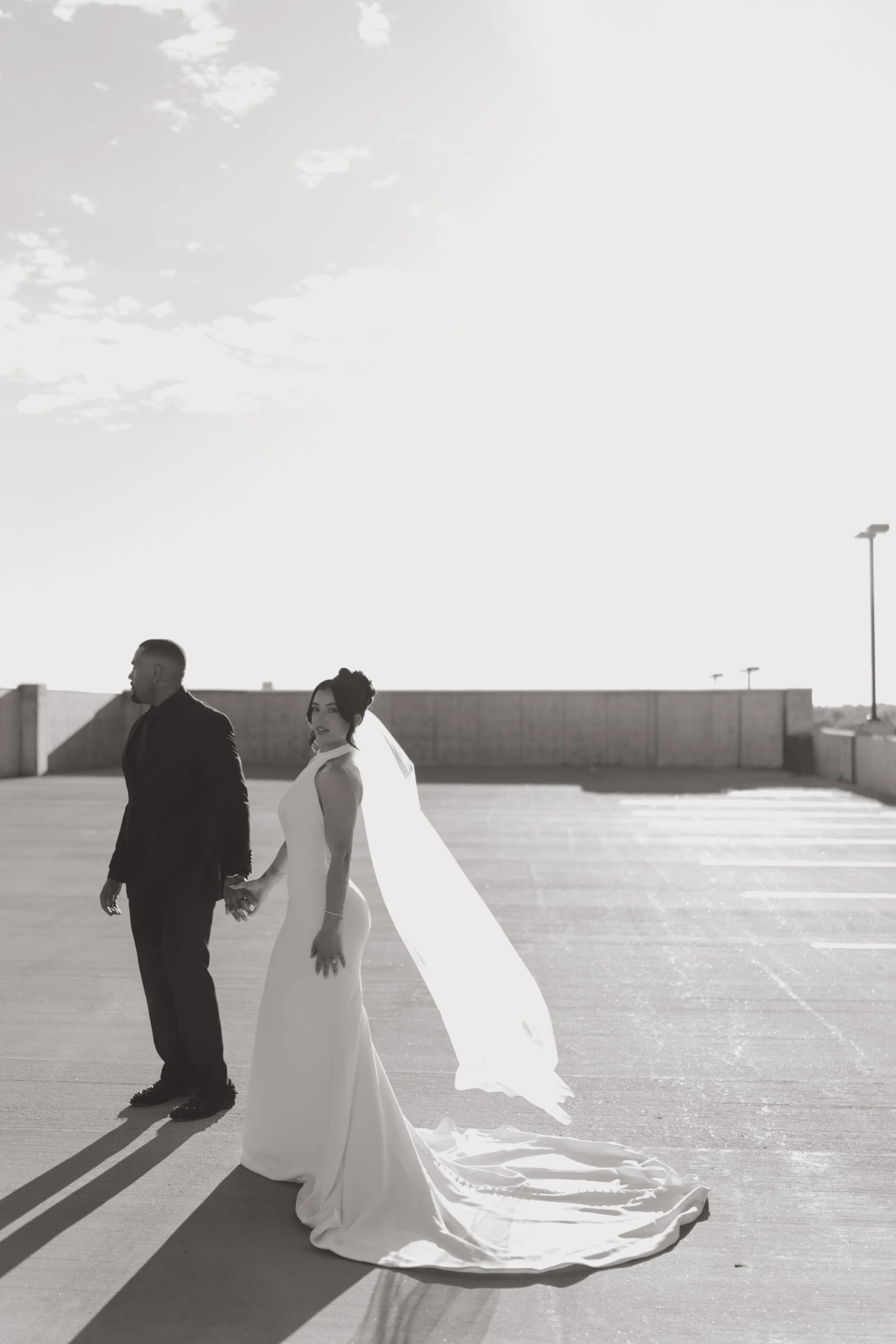 Black and white photo of a bride and groom standing hand in hand on a rooftop parking lot, with sunlight illuminating the scene and a clear sky in the background.