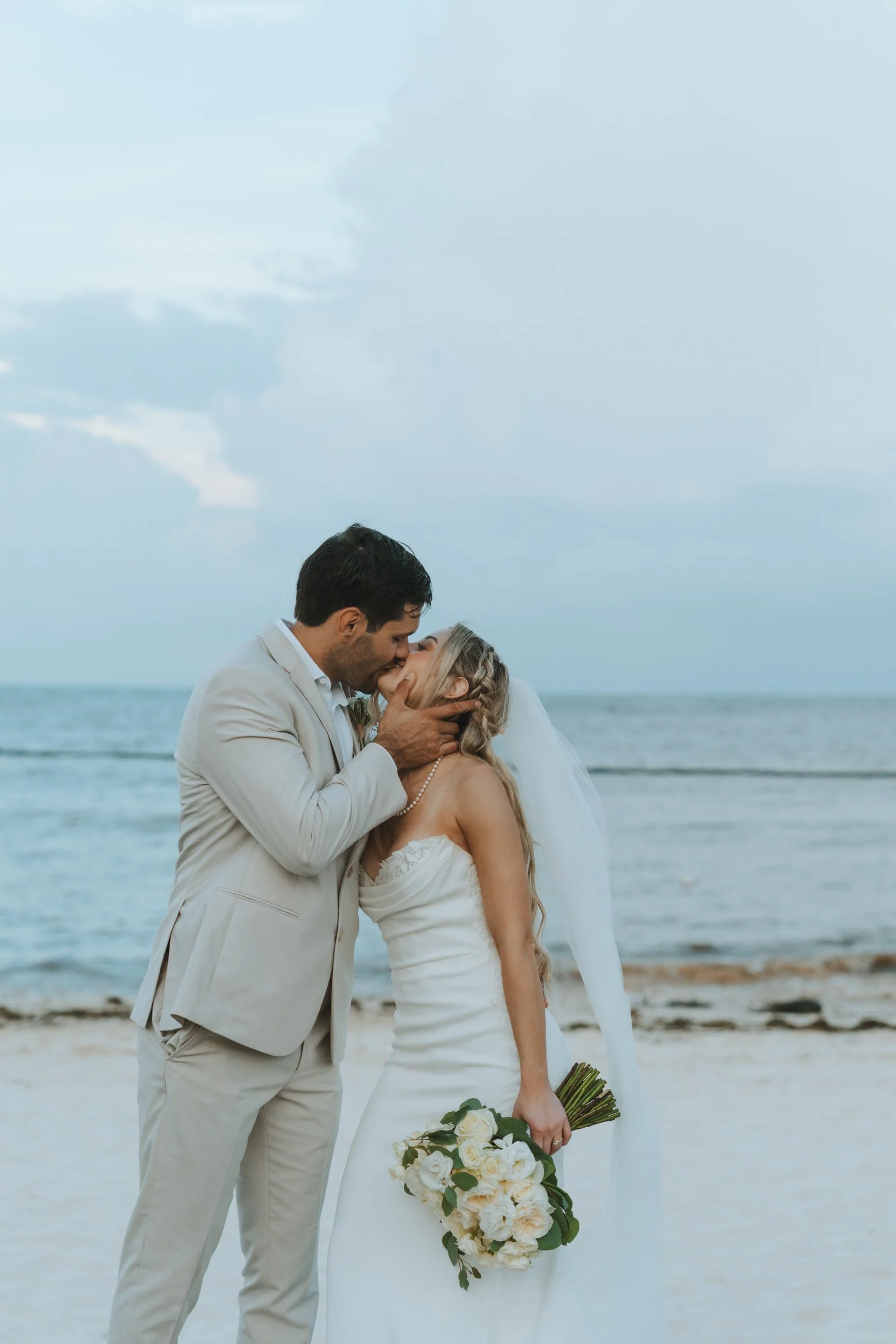 A bride and groom sharing a kiss on the beach, with the ocean and partly cloudy sky in the background.