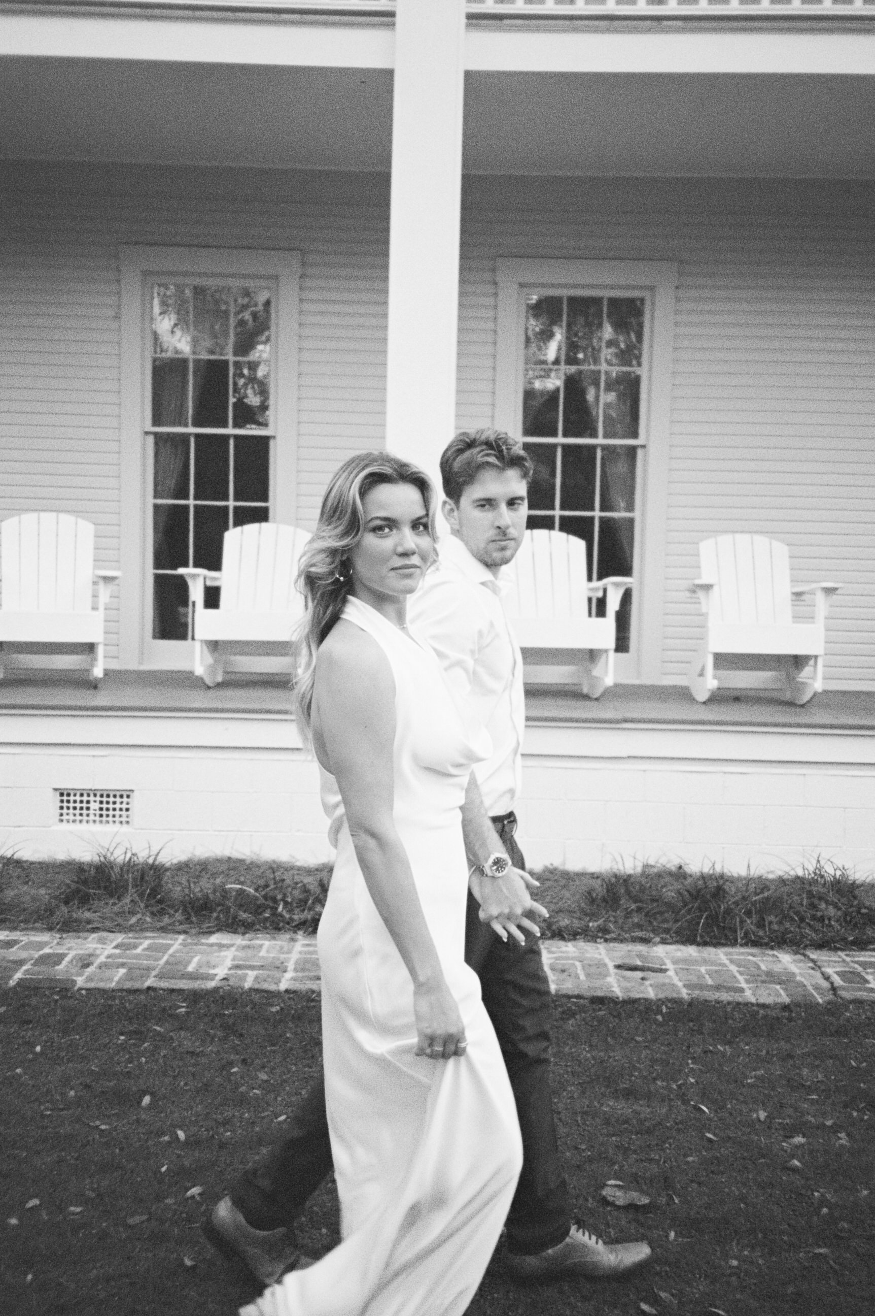 A black-and-white photo of a woman in a white dress and a man in dark trousers and a white shirt walking in front of a house with a porch featuring white chairs.