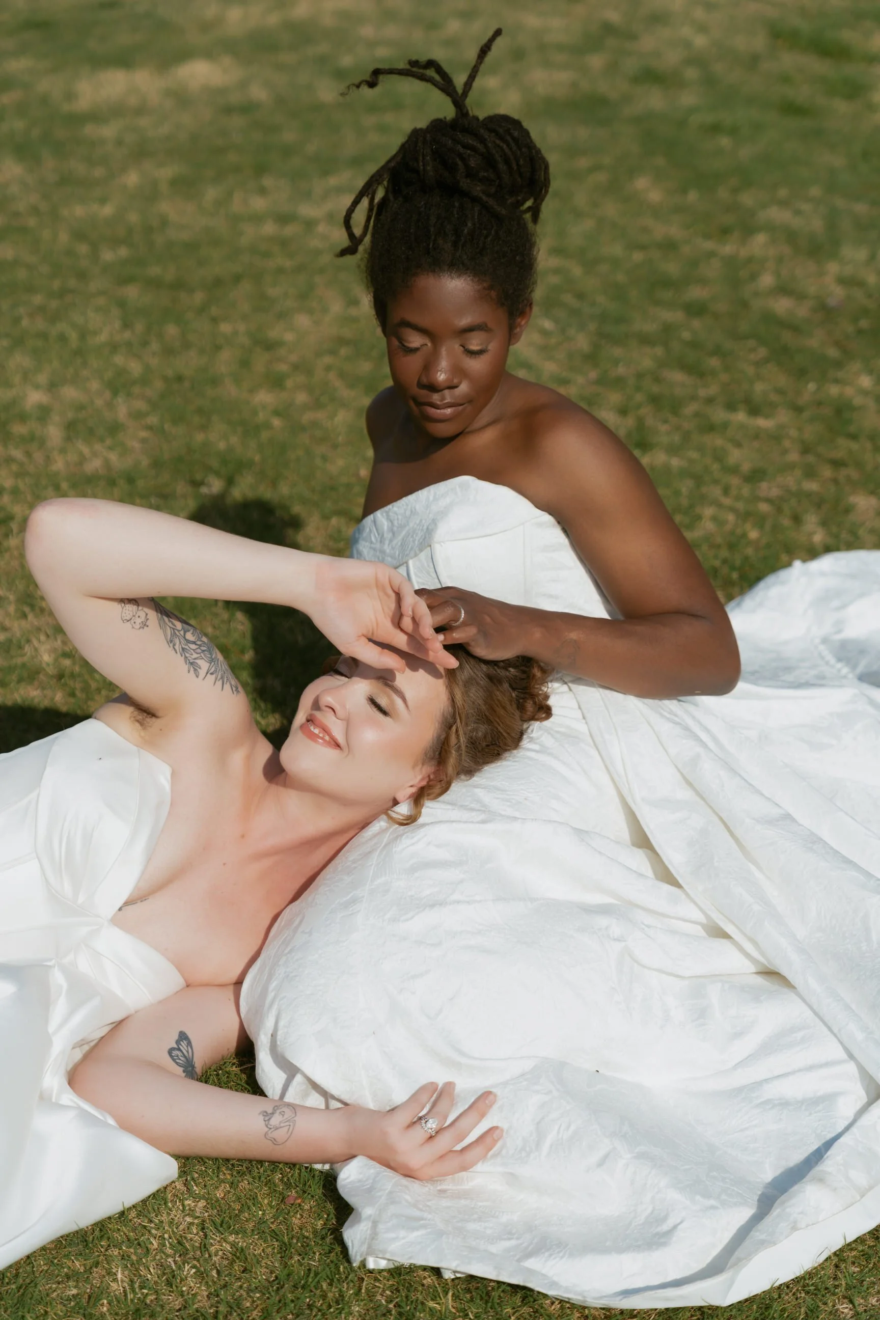 Two women in white dresses lying on green grass outdoors. One woman with dark skin and dreadlocks is sitting, while the other woman with light skin and tattoos on her arm is lying down and smiling.