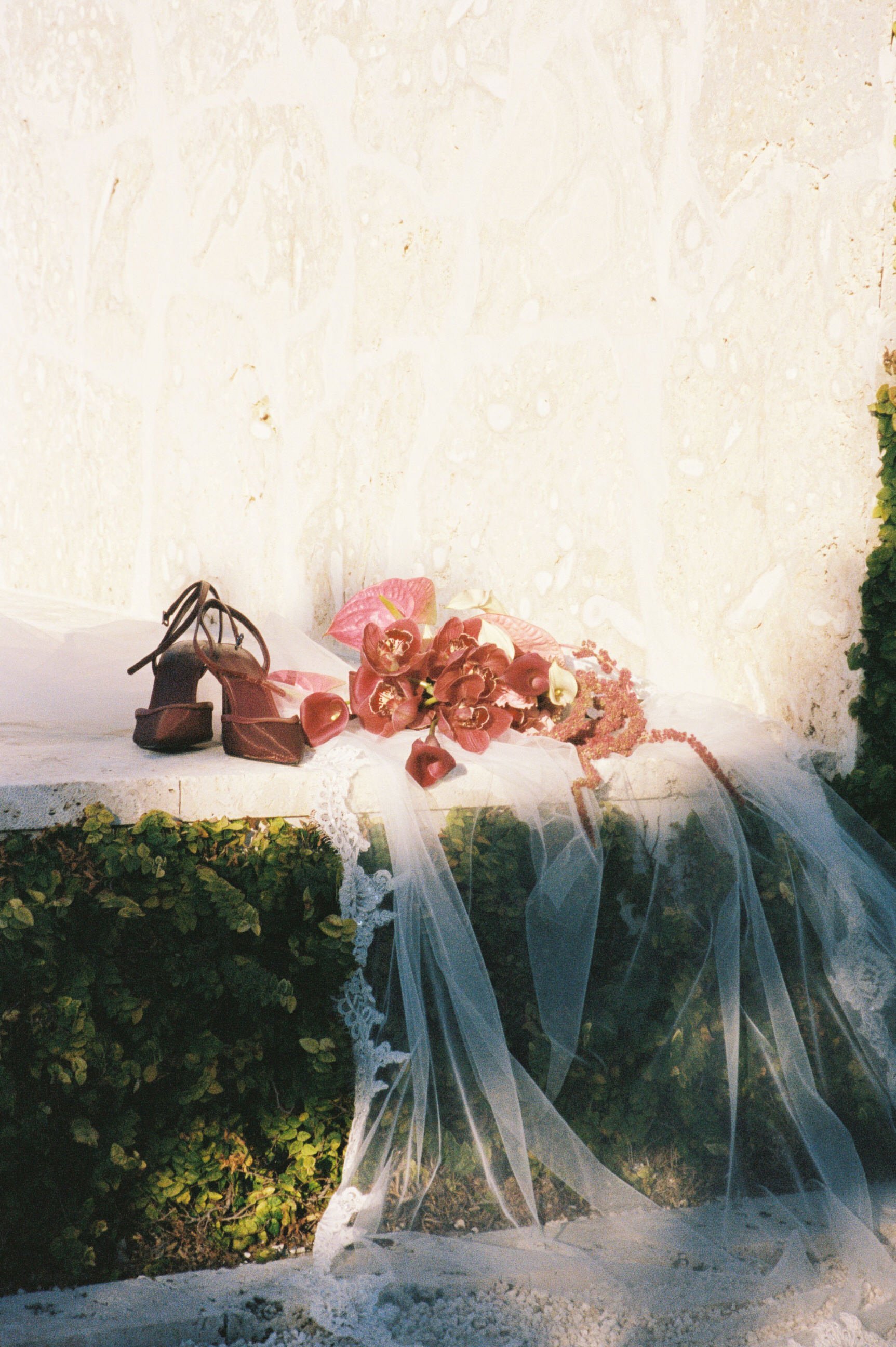 A floral arrangement with pink flowers, a pink veil, and a pair of high-heeled shoes on a white cloth on top of a green bush against a beige wall.