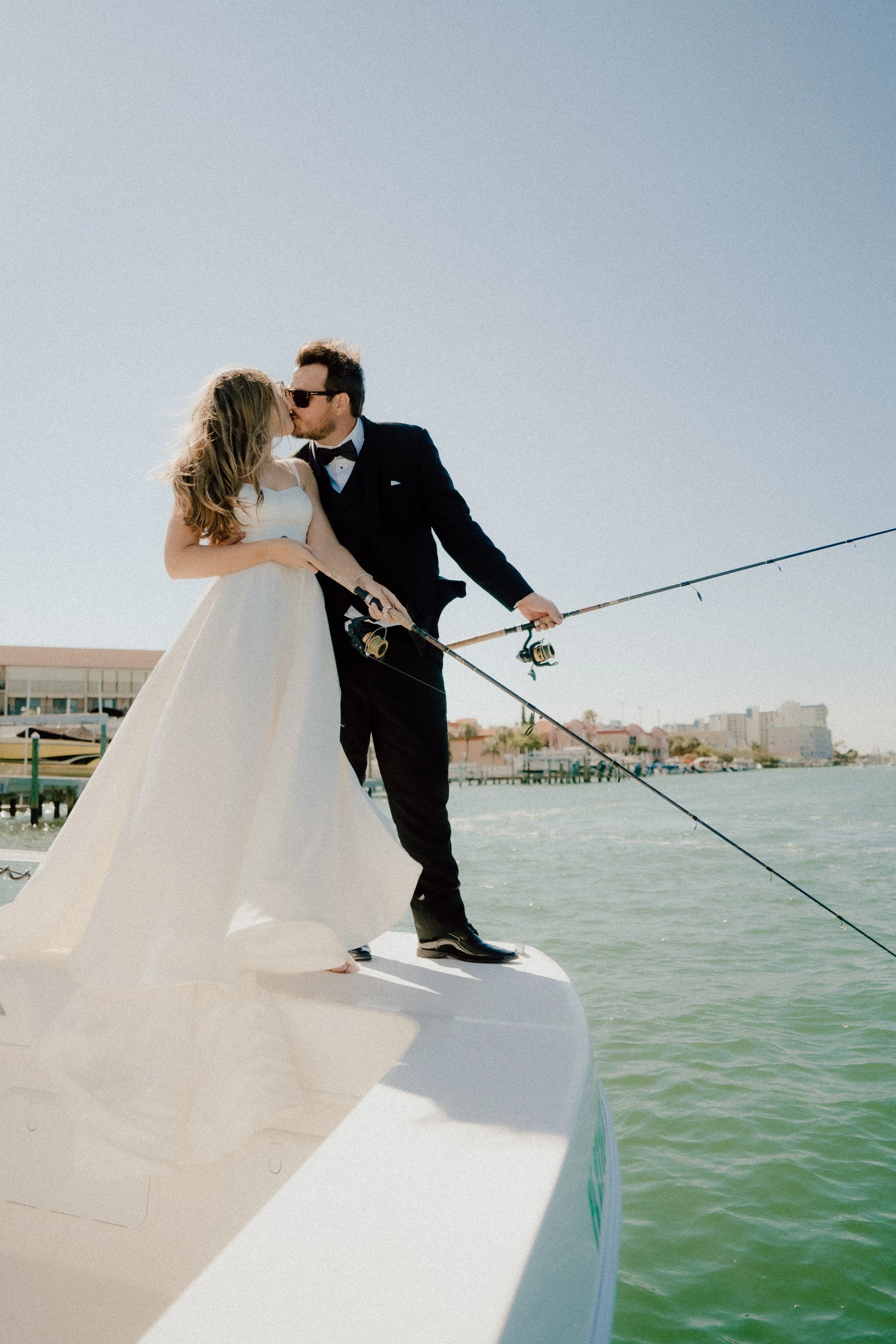 A bride and groom sharing a kiss on a boat, with the groom in a tuxedo and the bride in a white wedding dress, fishing rods in hand, near a waterfront with buildings in the background.