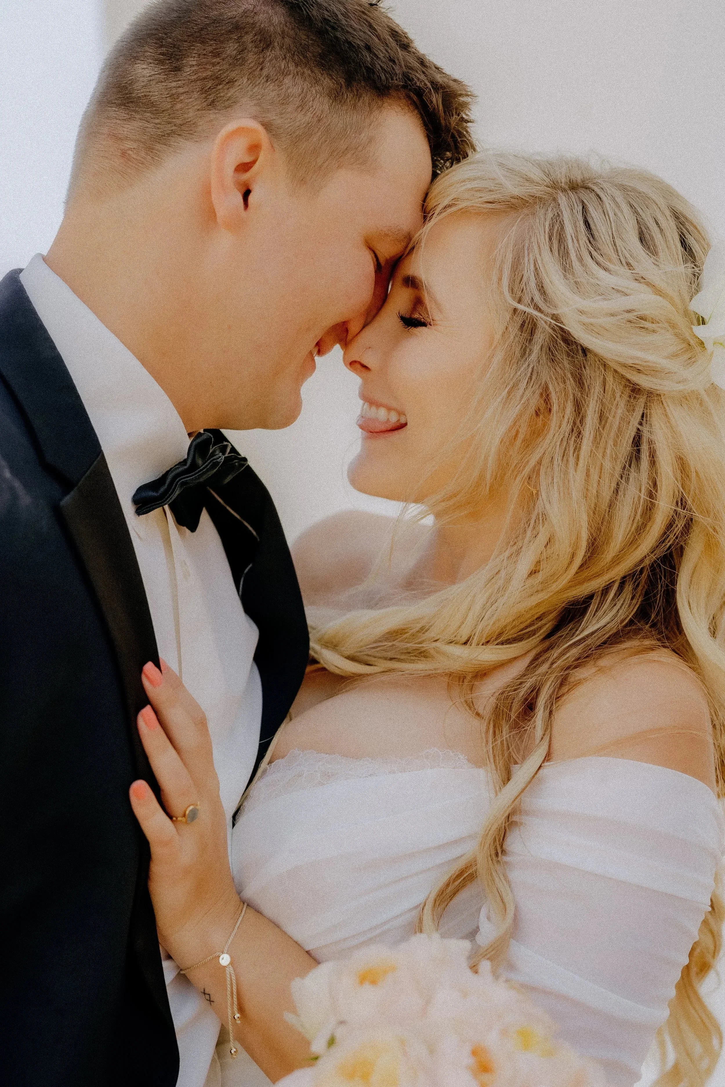 A couple is close together, smiling and touching foreheads. The man is in a tuxedo and the woman is in an off-shoulder wedding dress, holding a bouquet.