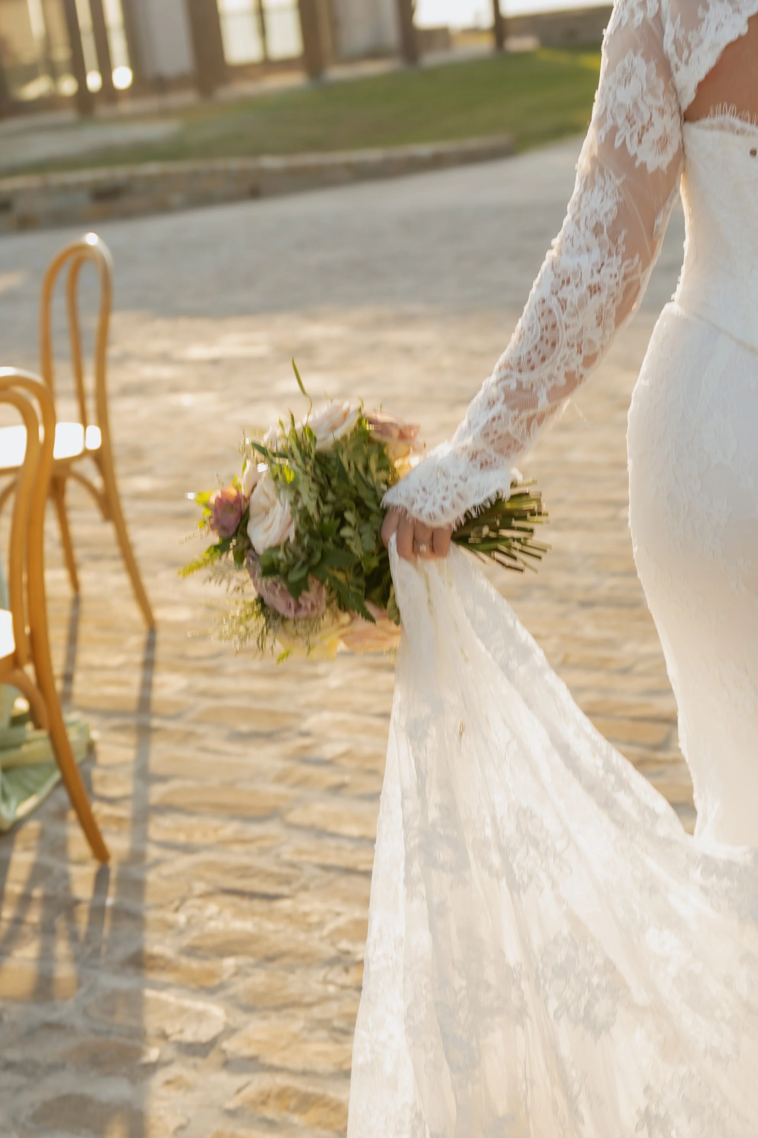 Close-up of a bride in a white lace wedding dress holding a bouquet of pink and white flowers outside during sunset.