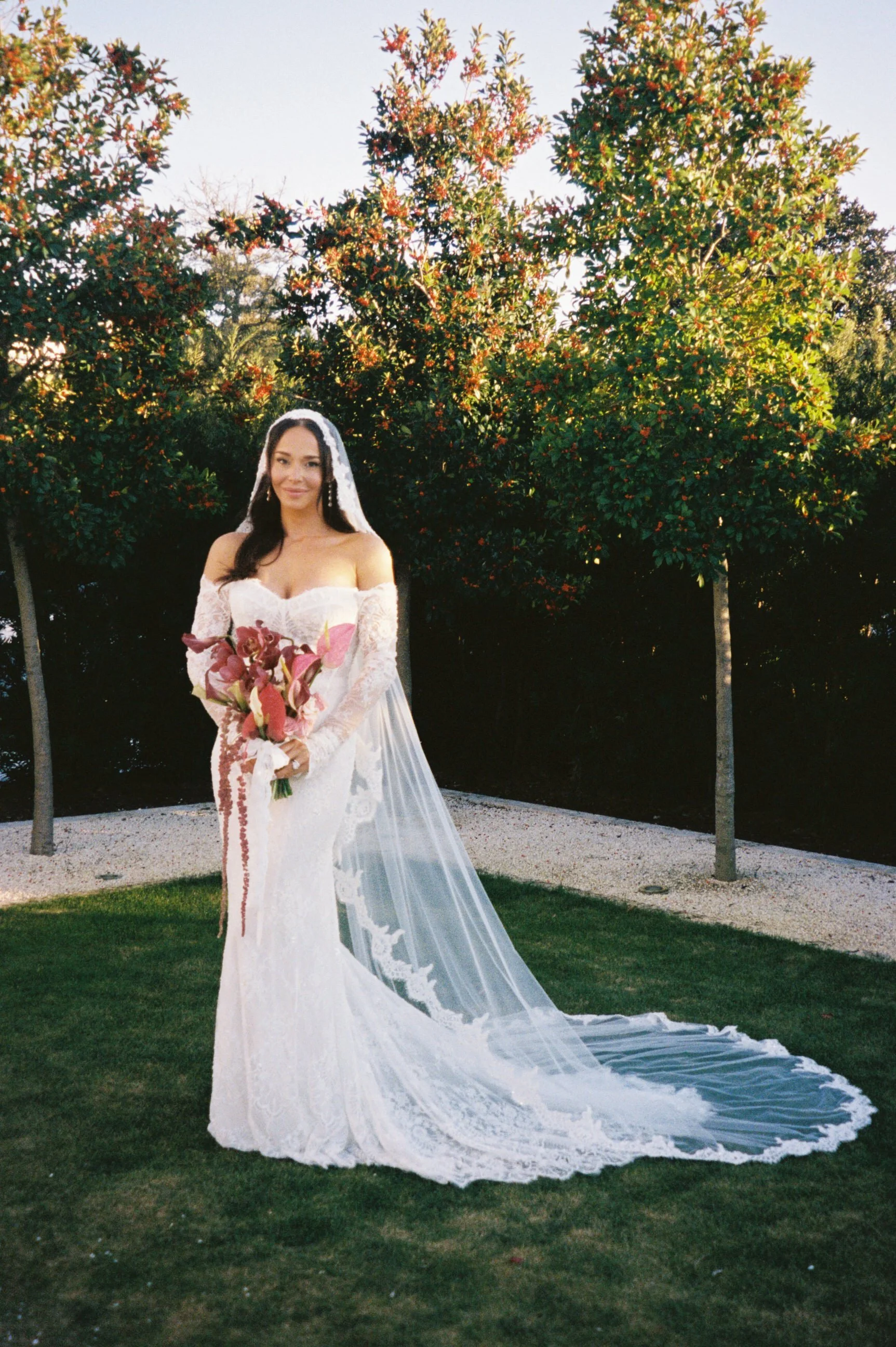 Woman in a white wedding gown holding a bouquet outdoors on a grassy area with trees in the background.