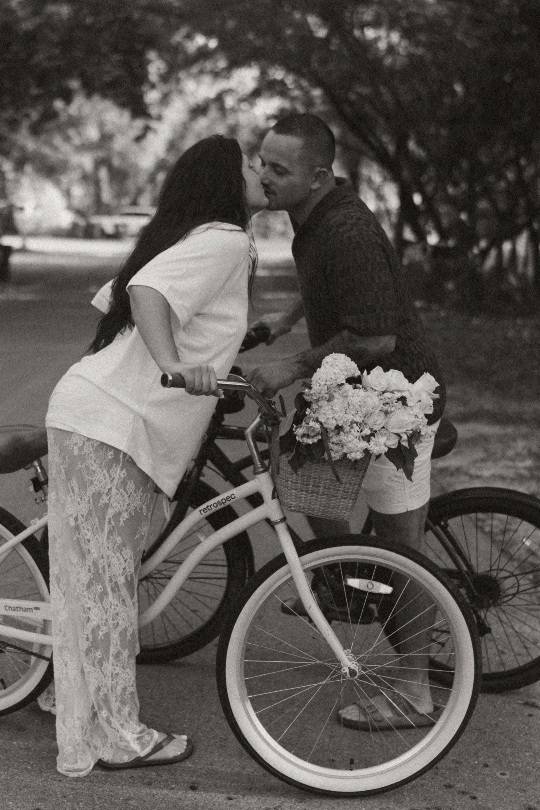 A couple sharing a kiss in front of bicycles, one with a basket of flowers, in a park-like setting.