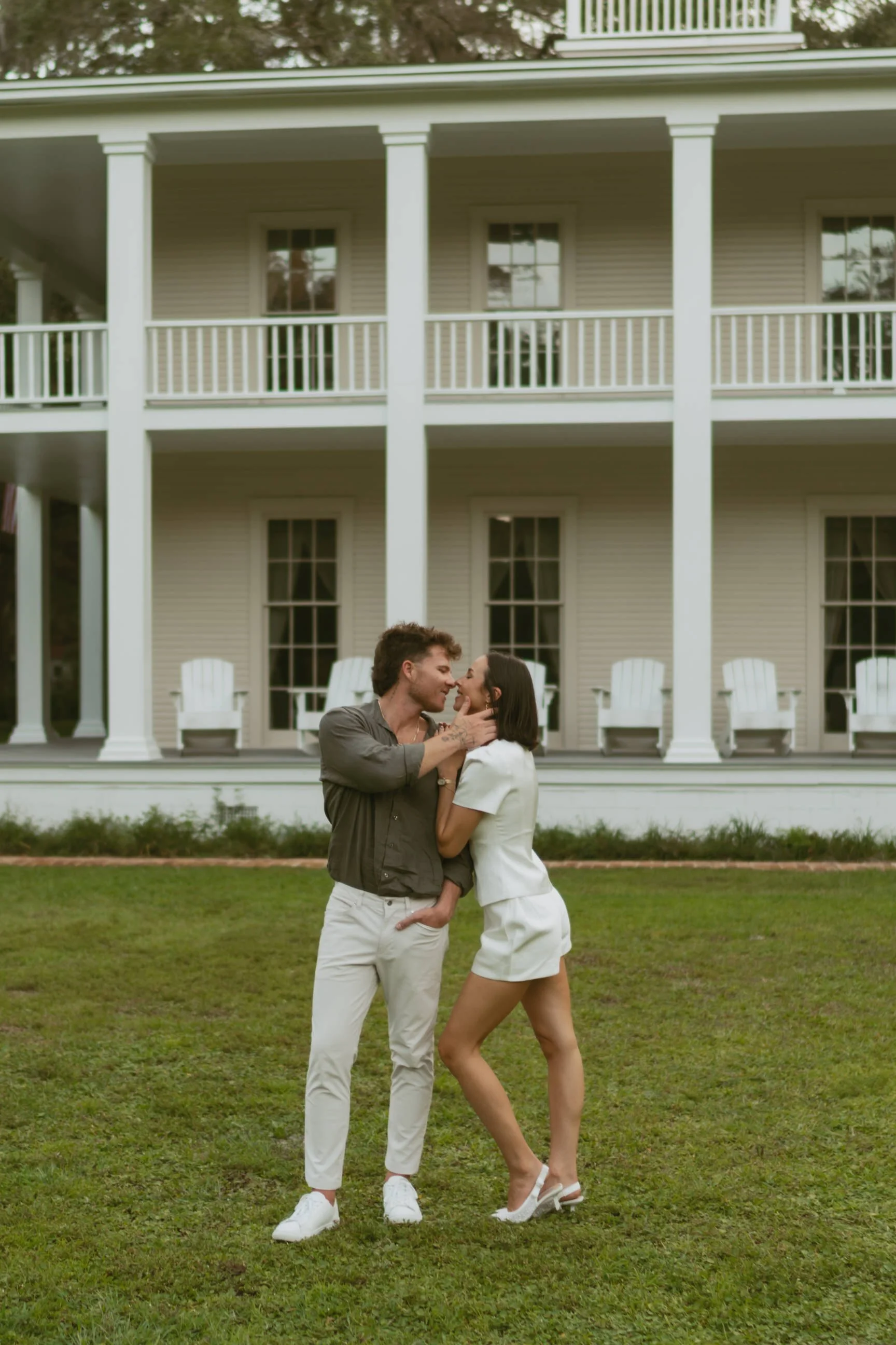 A couple sharing an intimate moment on a grassy lawn in front of a large, white, two-story house with a porch.