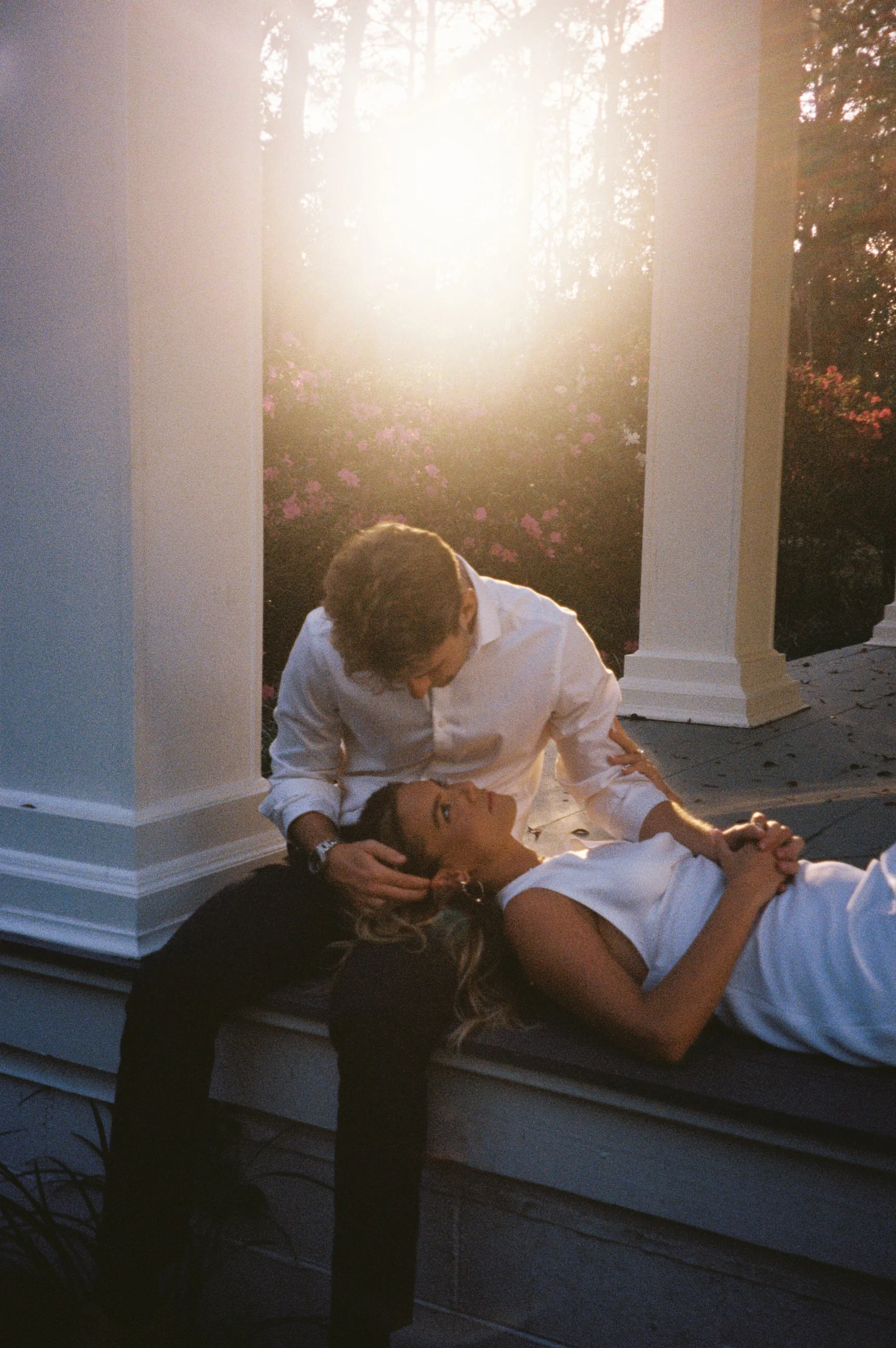 A man in a white shirt leans over a woman lying on a porch, with sunlight shining behind them and pink flowering bushes in the background.