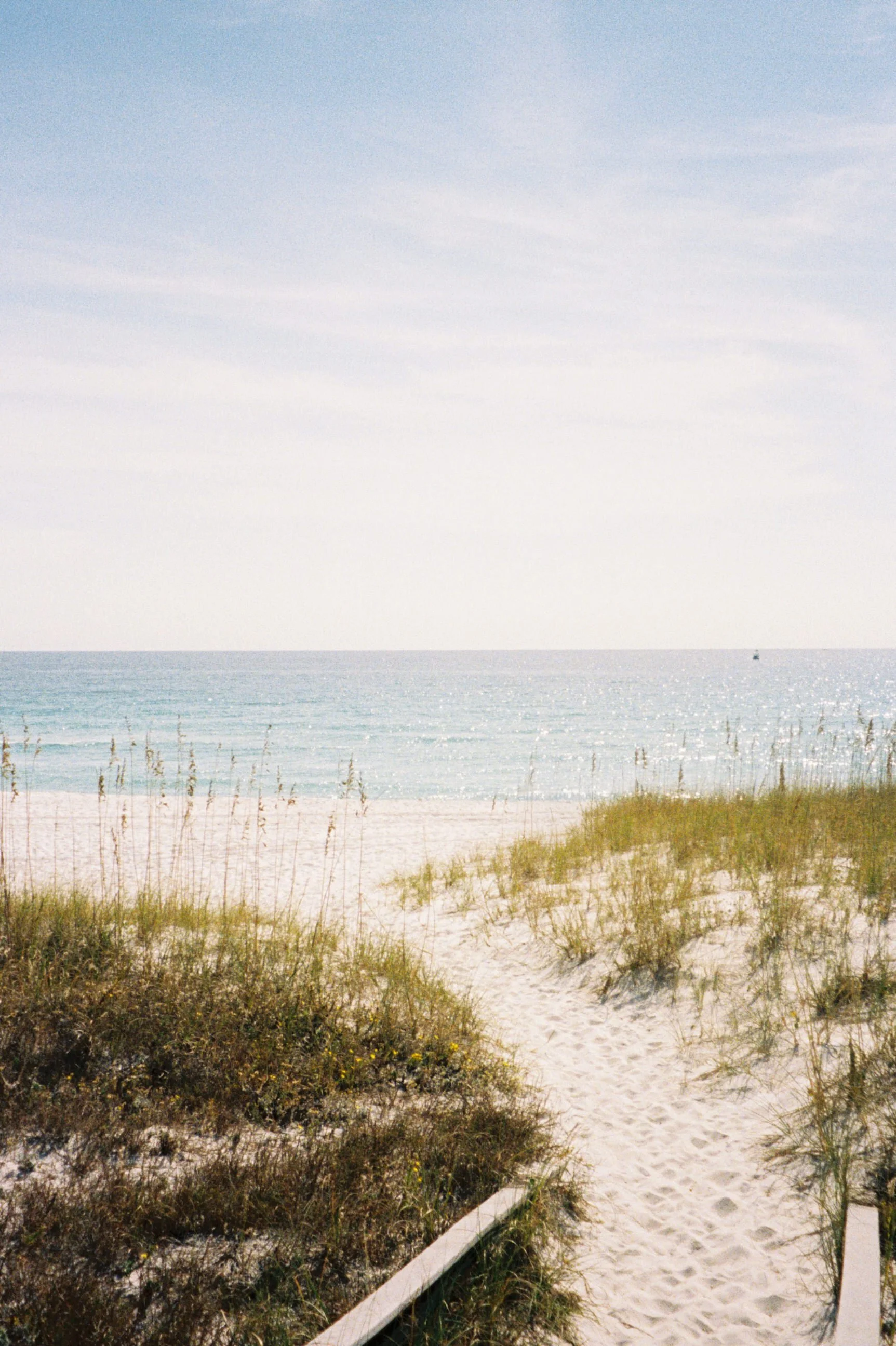 View of a sandy beach leading to the ocean with grassy dunes and a partly cloudy sky.